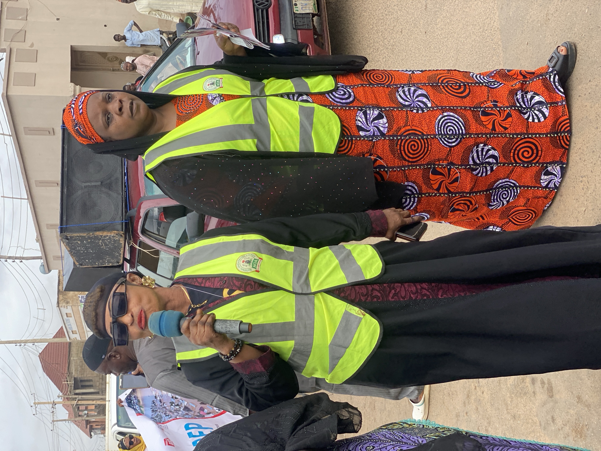 The image depicts a street scene in Rigachikun, Nigeria, where two women are central figures. The woman on the left, wearing a black dress, a black beret, and sunglasses, holds a microphone and is speaking. She is also wearing a bright yellow reflective vest over her dress, with a logo on it. The woman on the right, dressed in a black headscarf and a black open-fronted garment over an orange and white patterned dress, also wears a yellow reflective vest. She holds some papers in her hand. Both women are positioned in the foreground, facing towards the camera and appearing to be addressing an audience or participating in an event.
Behind them, a red vehicle is visible with a large speaker system mounted on top. Other people can be seen in the background, some in casual attire, and others wearing light blue and cream-colored clothing, suggesting a public gathering or procession. The environment appears to be an urban street with buildings and overhead power lines visible. The overall impression is of a public address or a community event taking place outdoors.