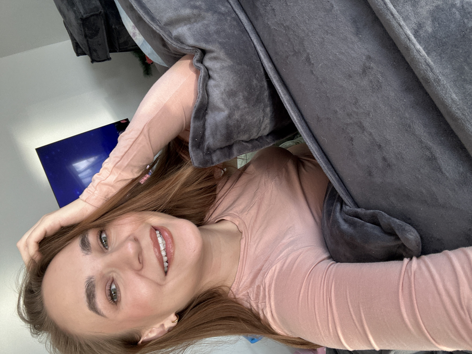 A fair-skinned woman with brown hair and green eyes smiles at the camera. She is wearing a pink long-sleeved shirt. Her right hand is on her head. She is being hugged by a large grey pillow. In the background, a television screen is visible on a white wall. A dark-colored curtain or drape hangs to the left of the screen. The scene is indoors. There is no information about this image being in the city of Kharnavy, Russia.