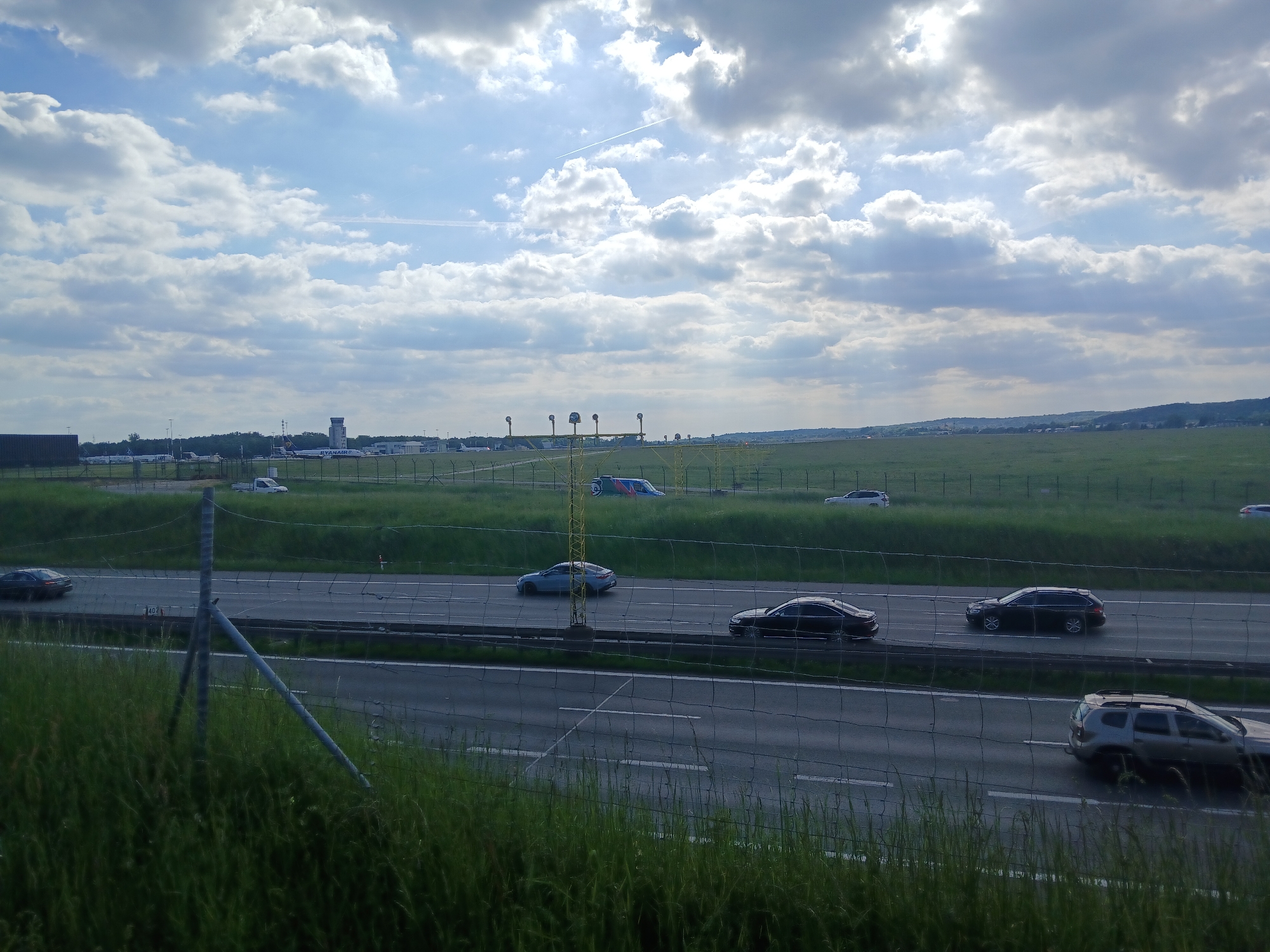 The image shows a multi-lane highway with several cars traveling on it, viewed from an elevated position through a fence. The highway is bordered by a grassy area. In the background is an airport with planes and a control tower visible. The sky is partly cloudy.