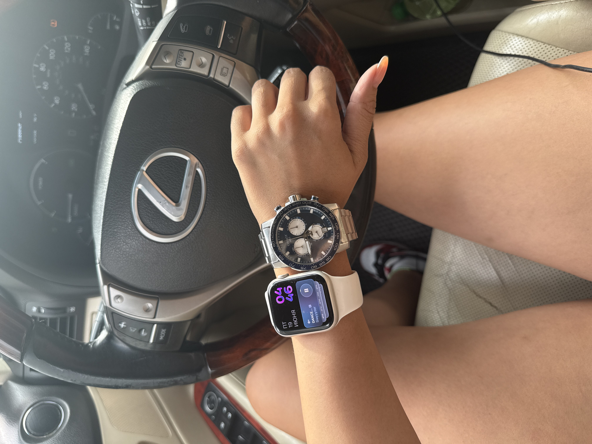 This image shows a person's hands and arms resting on a car's steering wheel. The person is wearing two watches, one a traditional analog watch with a silver band and blue face, and the other a white Apple Watch displaying the time "04:46", the date "19 ИЮНЯ" (June 19th), and "DANCE... SLAVYTEK". The background reveals parts of the car's interior, including the steering wheel with a Lexus logo, dashboard with a speedometer and other gauges, and a beige leather car seat. The person's legs are visible, one clad in a white, black, and red sneaker. The scene appears to be set during the daytime, with natural light illuminating the interior. There are no explicit weather cues, but the overall lighting suggests it's not overcast. The location is identified as Almaty, Kazakhstan. The image captures a moment of stillness, possibly while driving or stopped.
