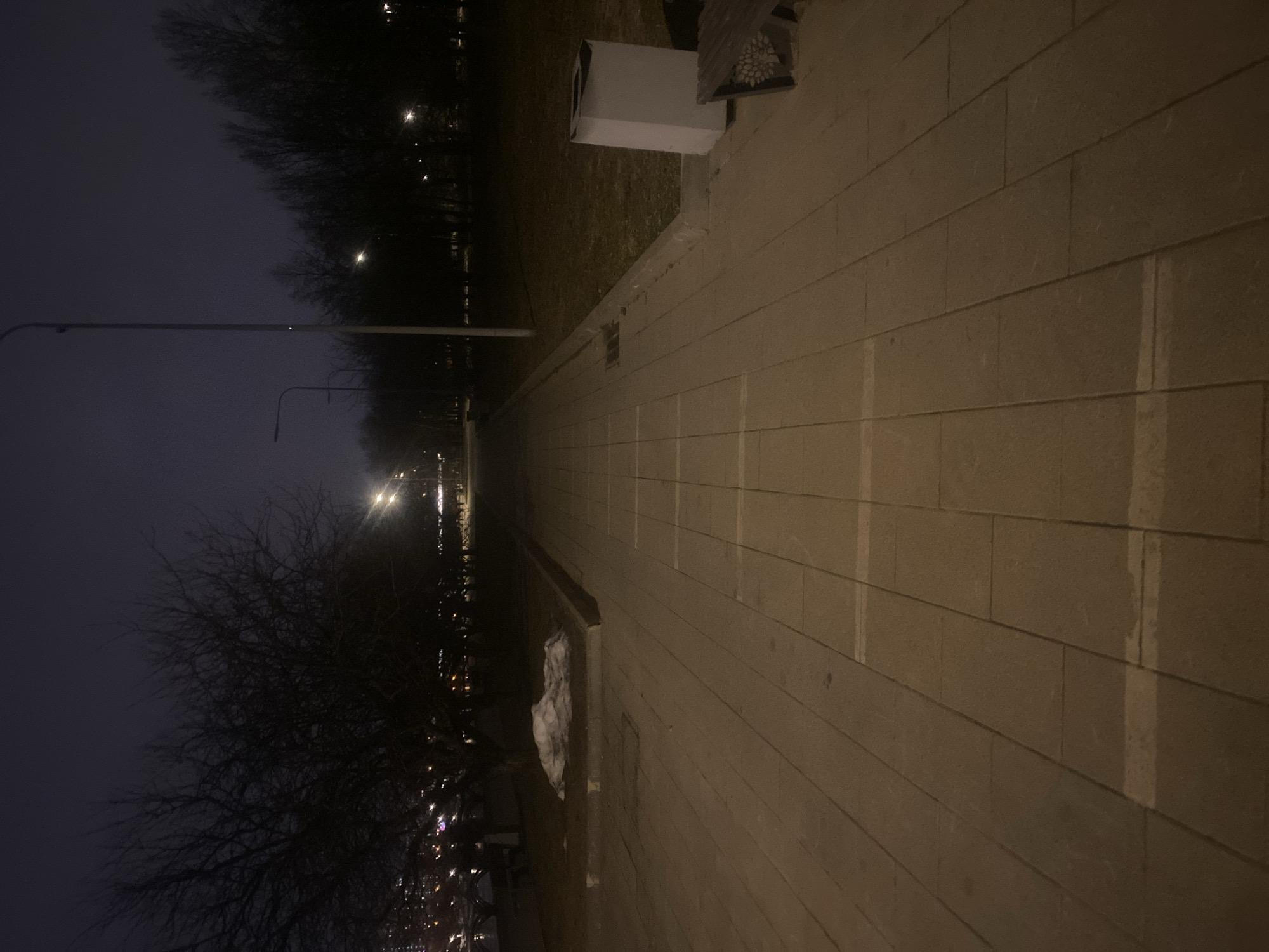The image shows an outdoor walkway at night. The walkway is paved with rectangular stones. To the left, there are trees and streetlights. The overall illumination is dim, and the sky appears dark. There is a white object, possibly a trash can, visible on the right side of the walkway. At the end of the path, there are more lights.