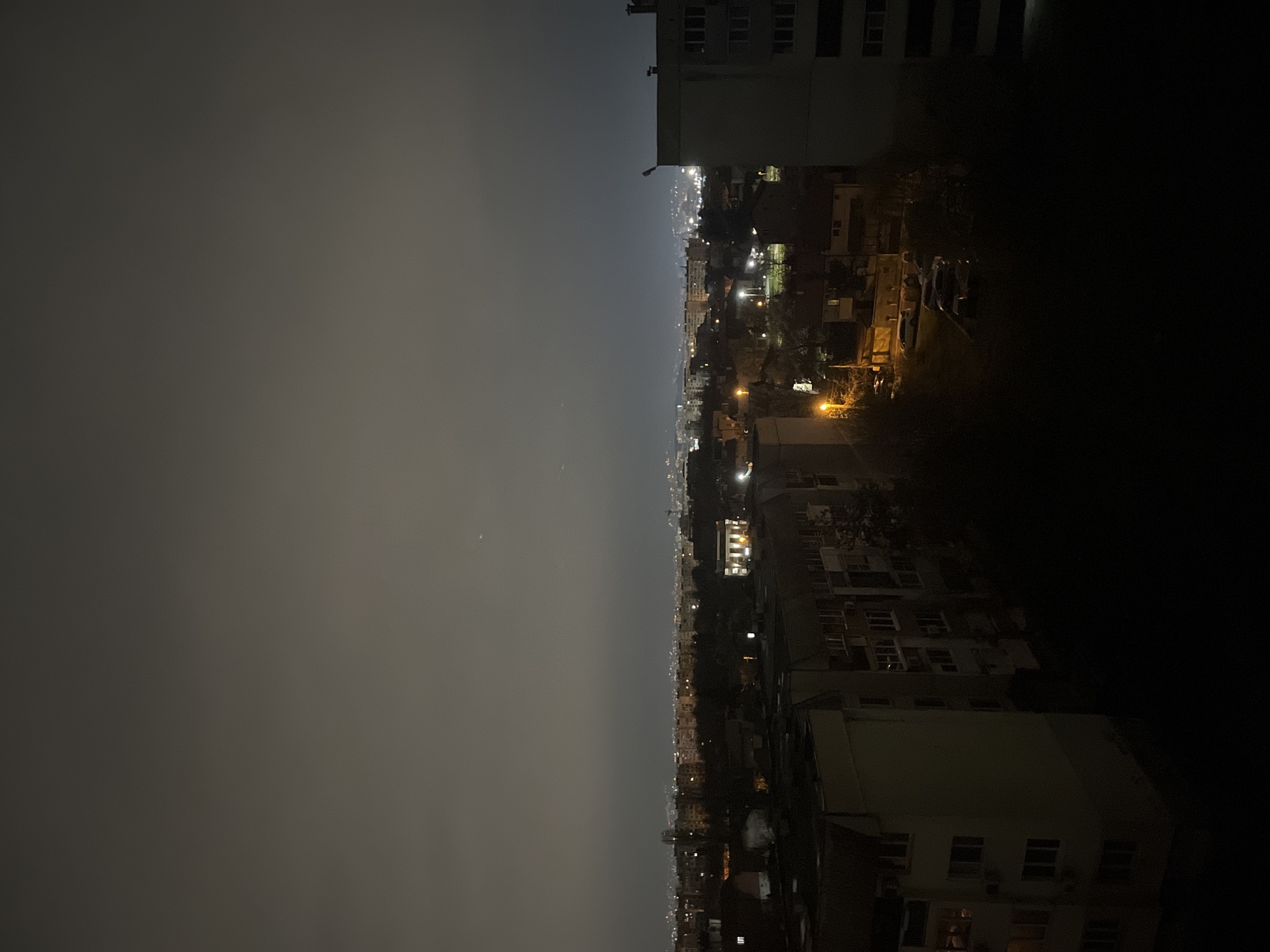 The image depicts a night view of Sofia, Bulgaria, from an elevated perspective. The scene is dominated by a city skyline with numerous buildings of varying heights, illuminated by streetlights and internal building lights. A heavy overcast sky obscures the upper portion of the frame. The lower part of the image is enveloped in darkness, obscuring ground-level details. The overall atmosphere is one of subdued urban activity under a cloak of nighttime and cloud cover.