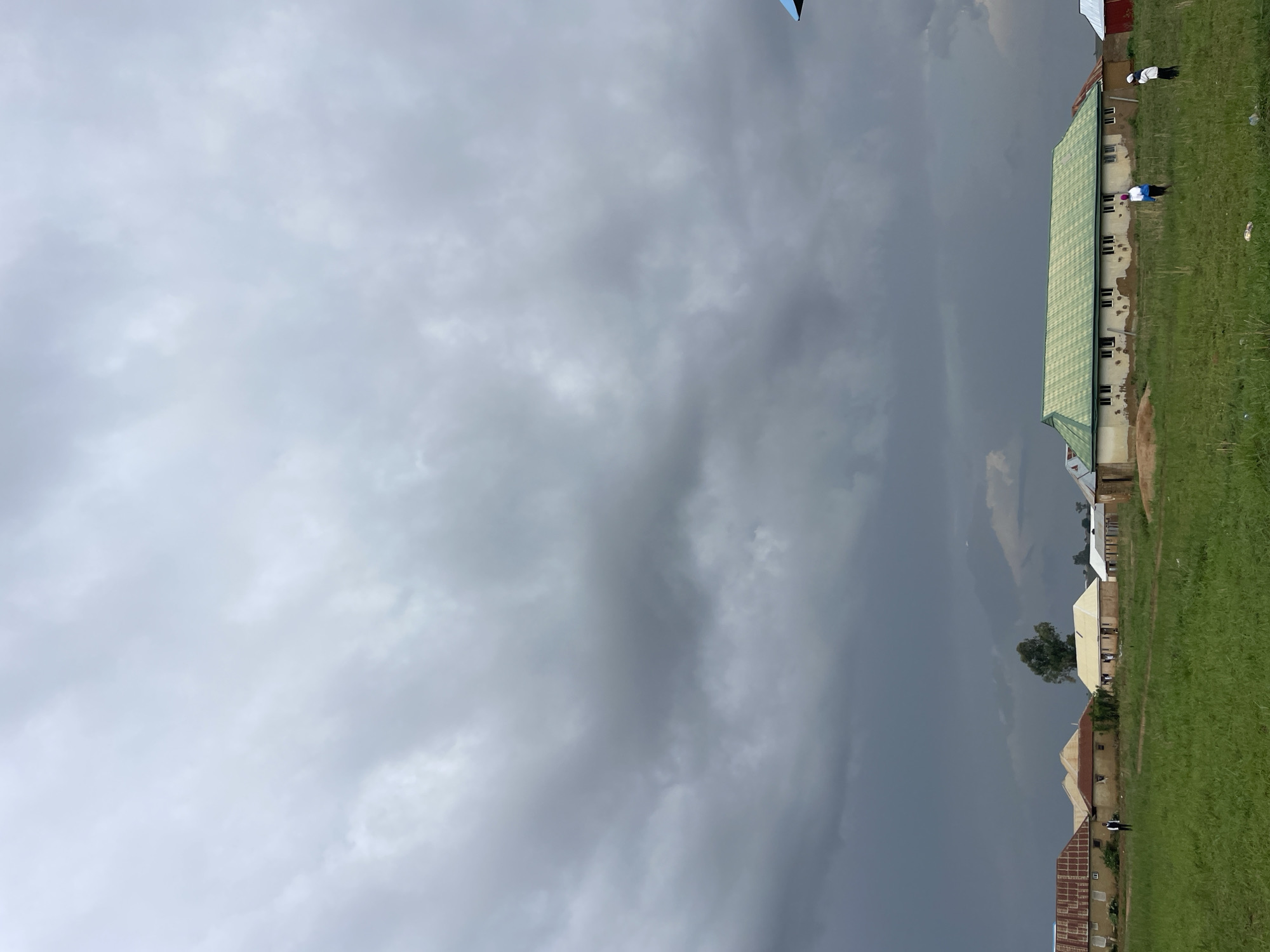 The image depicts a view of Durimi, Nigeria under a heavily clouded sky. A row of buildings with varying roof colors (red, green, silver) lines the right side of the frame, bordered by green grass. Two individuals are visible near the building with the green roof; one wears white and black attire, and the other wears a blue head covering with white and black attire.