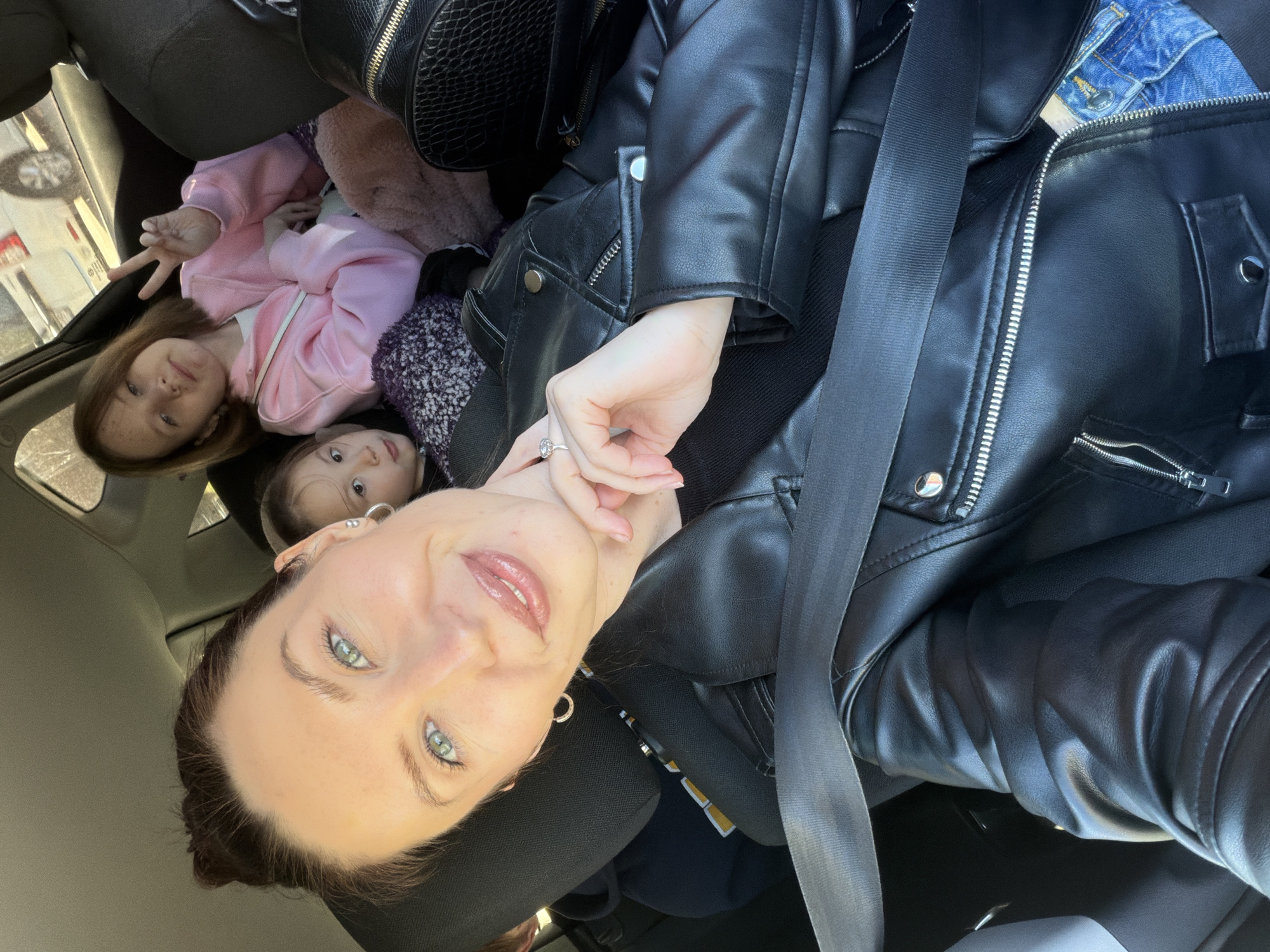 The image shows a selfie taken inside a car. A woman wearing a black leather jacket is facing the camera, with a seatbelt across her chest. In the background, two young girls are lying in the backseat. One girl is wearing a pink jacket and making a peace sign with her fingers. The other girl is partially visible behind her. A black purse is visible above the girls. The location is implied to be in Perm, Russia.