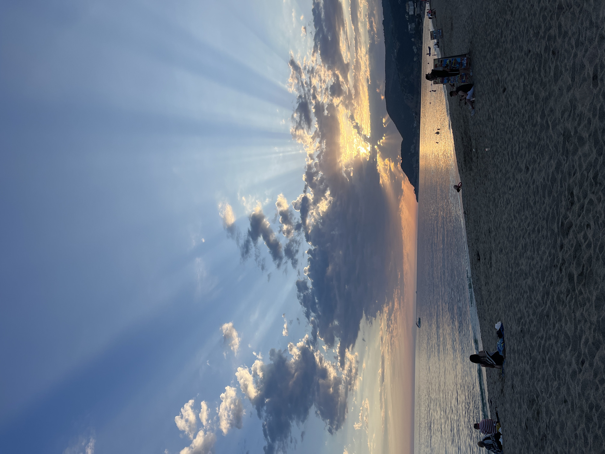The image shows a beach scene in Alanya, Turkey, at sunset. Sunlight streams through clouds, creating crepuscular rays in the sky. The sea reflects the golden light. The beach is populated with people, some sitting and relaxing. The sand is dark, likely due to the time of day. A dark, mountainous shape is visible along the coastline in the distance.