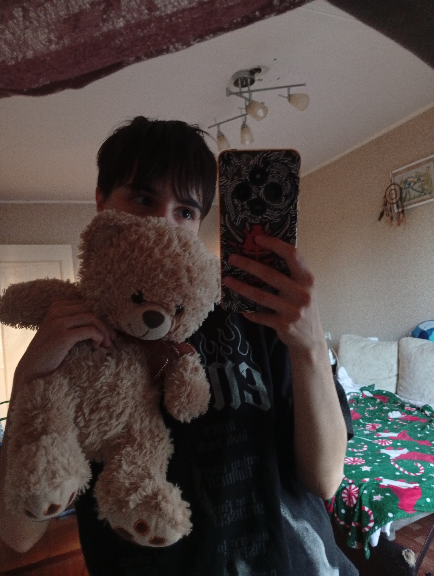 A person holds a teddy bear in front of their face while taking a selfie with a smartphone. The person has dark hair and is wearing a black t-shirt. The phone has a black case with a dragon design. The teddy bear is light brown with a brown ribbon around its neck. The background shows a room with light-colored walls, a bed with a Christmas-themed blanket, and a ceiling light. A dreamcatcher hangs on the wall. A partial view of a mirror is visible in the top left. The exact location in Naberezhnye Chelny, Russia, is impossible to determine from the image.