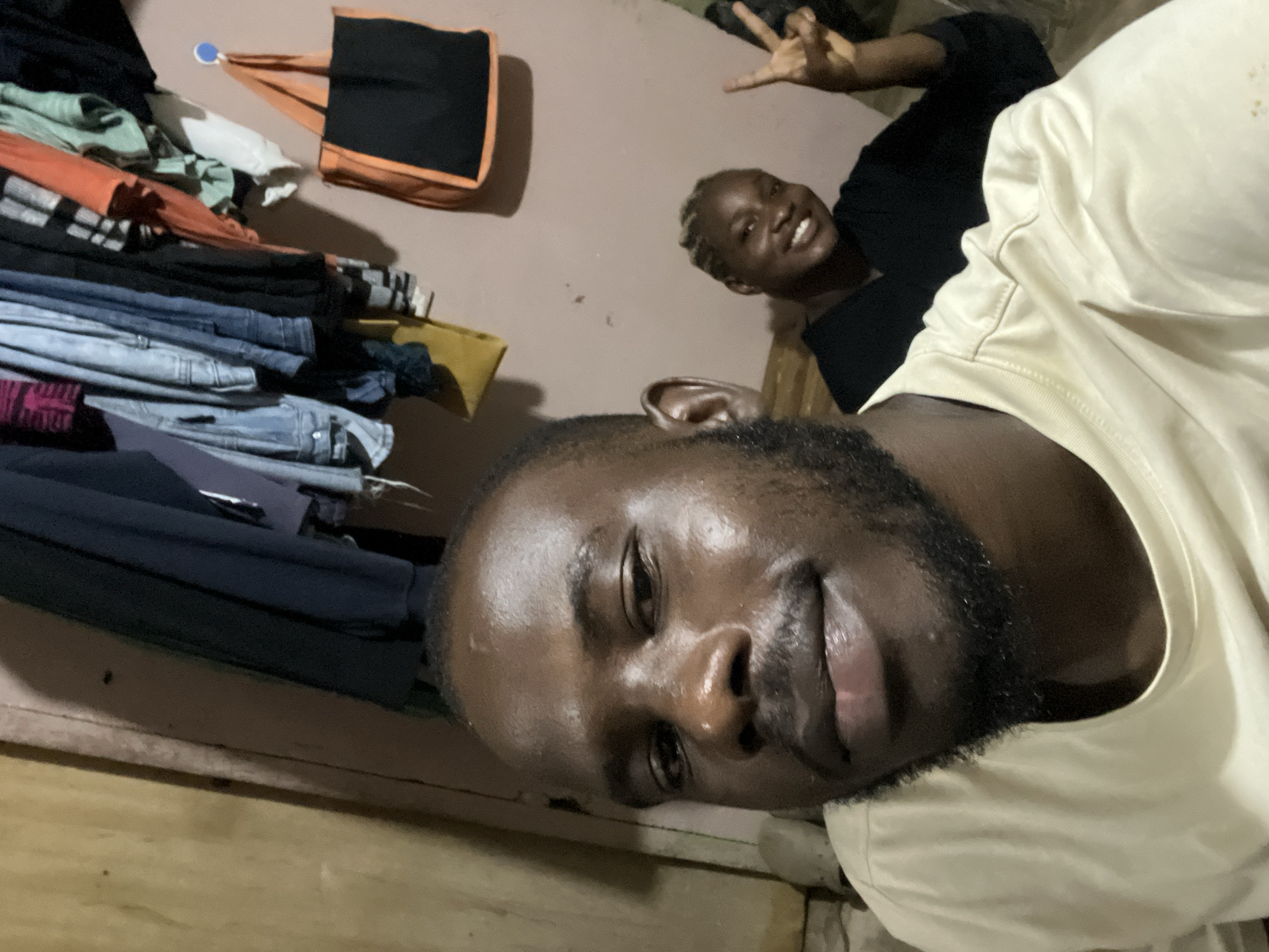 In Rafin Par, Nigeria, a man and a woman pose for a selfie indoors. The man is in the foreground, his face occupying the left side of the frame. He has dark skin, short hair, and a beard, and is wearing a light-colored t-shirt. He is looking directly at the camera with a slight smile. Behind him and to the right, the woman is visible, smiling and making a peace sign with her fingers. She has braided hair and is wearing a dark top. The background shows a wall with clothes hanging on it, possibly in a closet or on a clothesline. A black and orange bag hangs on a hook on the wall. The lighting suggests it is likely evening or indoors.
