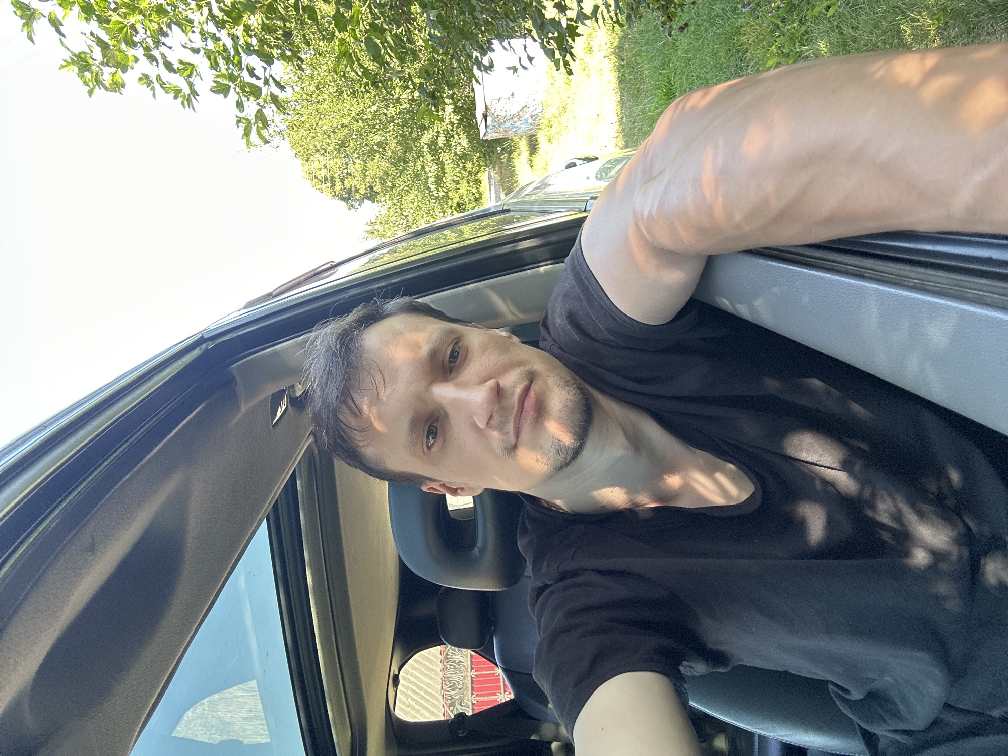 A man is sitting in the driver's seat of a car, with his left arm resting on the open car door. He is wearing a black t-shirt and has short, dark hair and stubble. Sunlight filters through the trees and casts dappled shadows on him and the car's interior. The car's sunroof is open, revealing a clear blue sky. In the background, lush green trees and a fence are visible. The overall setting suggests a rural or suburban environment, possibly in the vicinity of Shatava, Ukraine, given the vegetation and the style of the fence.