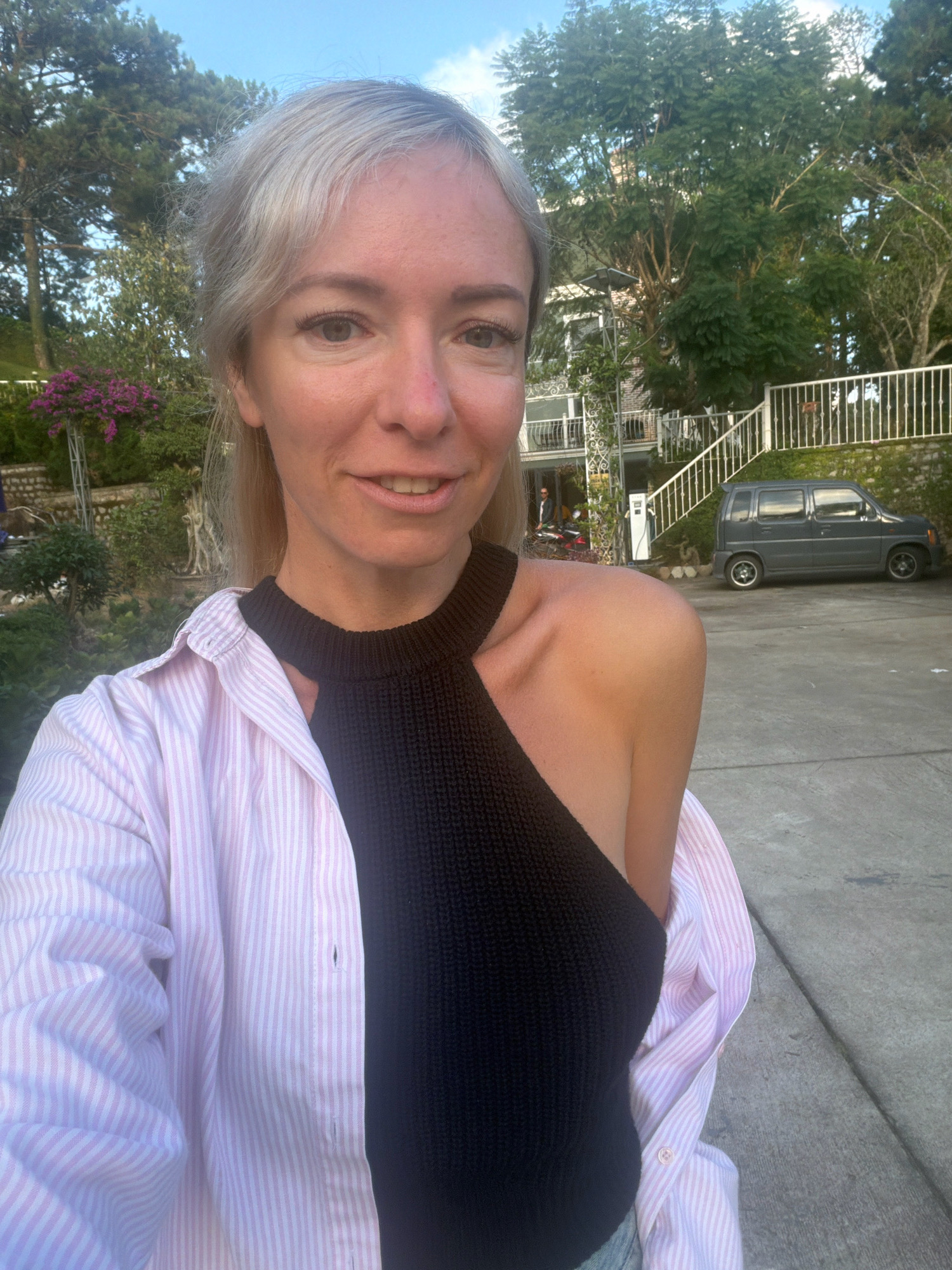 In a selfie taken outdoors, a woman with blonde hair is captured from the chest up. She is wearing a light pink and white striped button-down shirt, unbuttoned, revealing a black halter top. Her left shoulder and upper arm are visible. The background shows a paved area, a grey car parked to the right, a white staircase leading to a building with balconies, and lush green trees. In the distance, a person on a motorcycle is visible near the building entrance. The sky is a clear blue. The image appears to have been taken in the city of Xuân Trường - Đà Lạt, Vietnam, given the architecture and vegetation.
