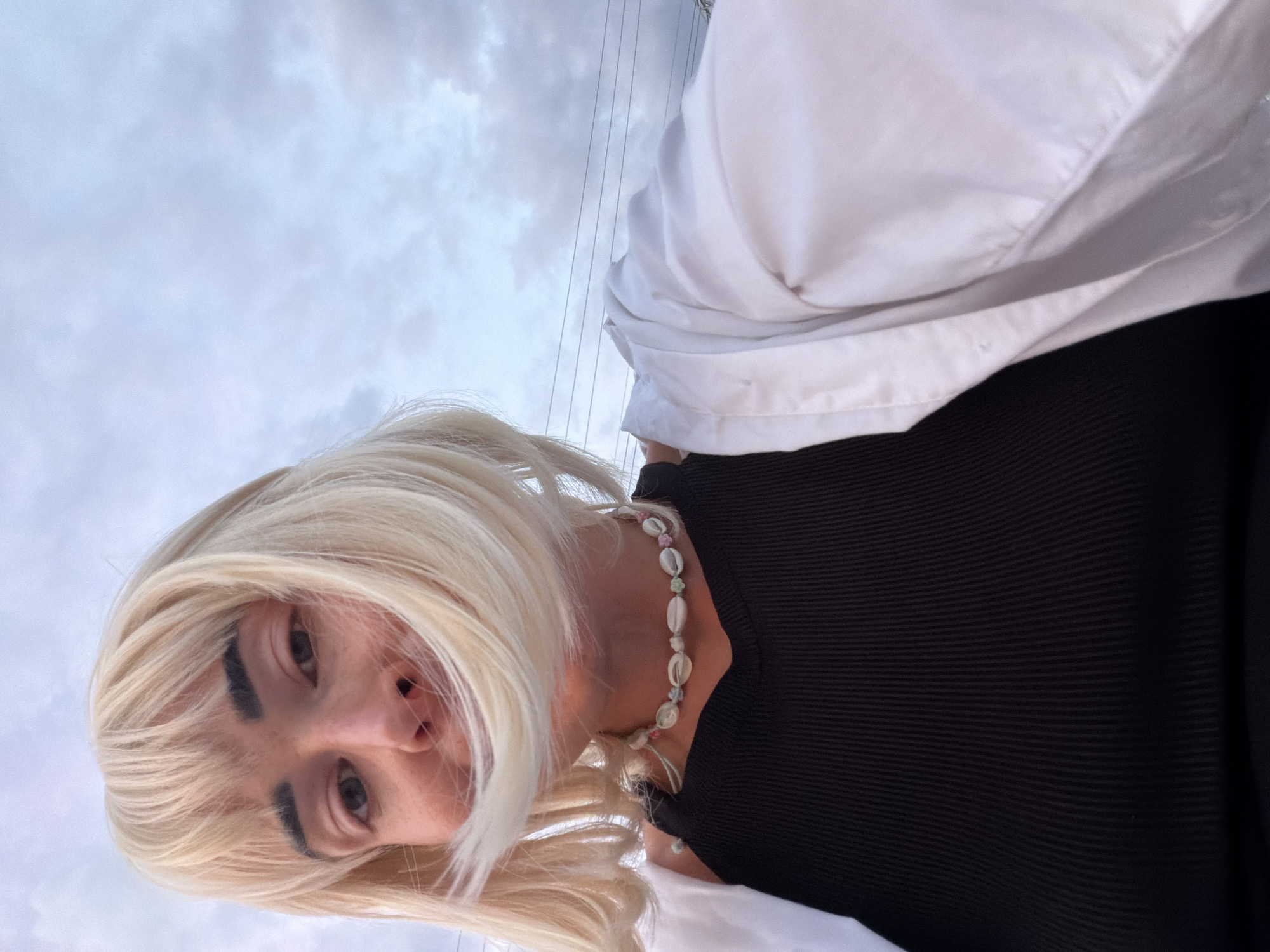 In a close-up, low-angle selfie, a person with blonde hair and bangs is captured against a cloudy sky. Their face is partially obscured by their hair, which is being blown by the wind. They are wearing a black ribbed tank top and a white unbuttoned shirt over it. A shell necklace with colorful beads adorns their neck. The background shows a pale sky with scattered clouds and power lines. The overall setting suggests an outdoor environment, possibly in a city, though the exact location is unknown.