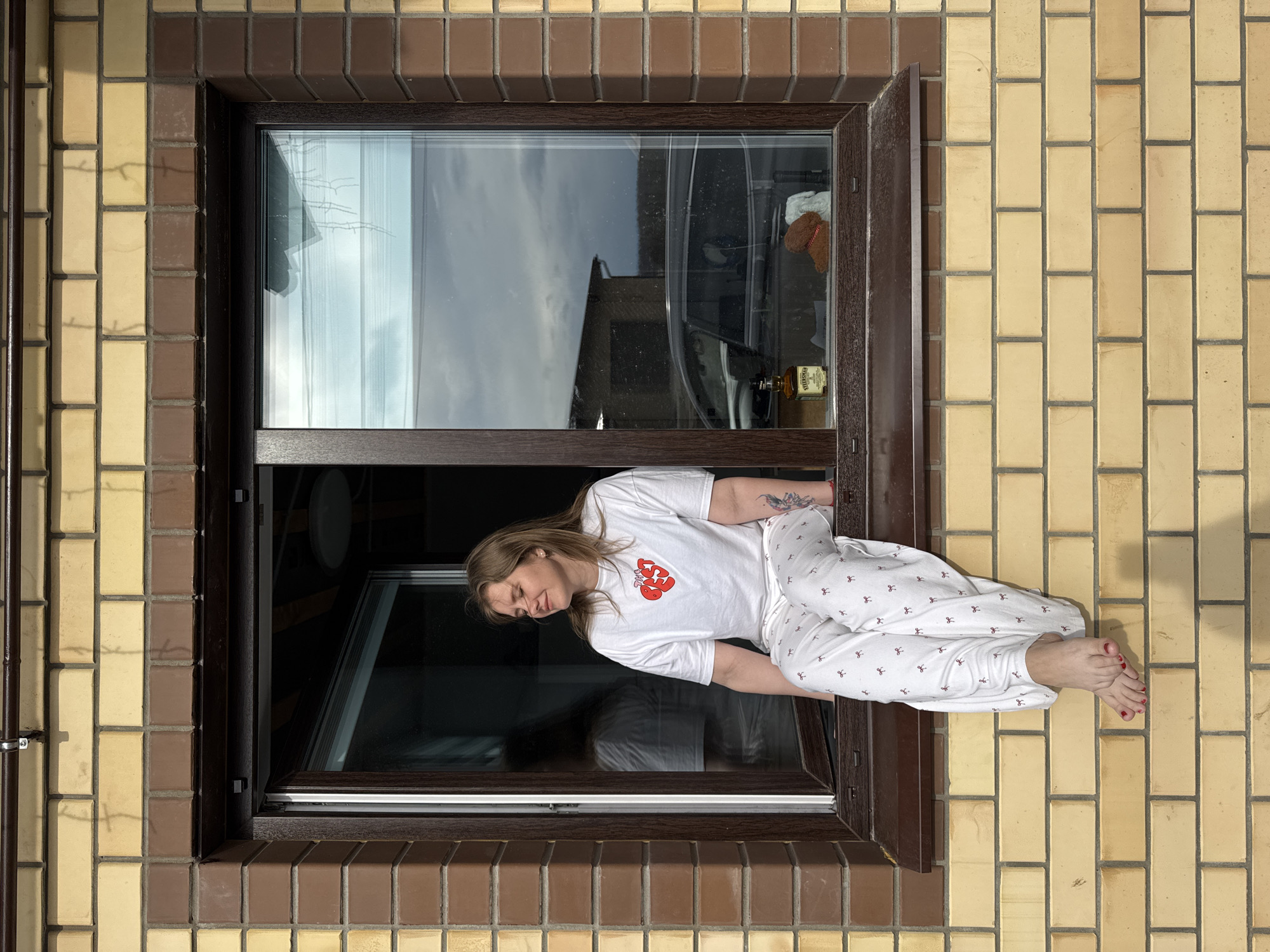 A medium, eye-level shot captures a light-skinned female individual positioned in the open window of a brick building. She is wearing white pajamas with a pattern and a white t-shirt. She is leaning towards the inside of the building and her body is partially outside of the window. Inside the building, a liquor bottle, a white cloth, and another building can be seen reflected in the glass. The building's exterior is constructed from yellow and brown bricks. The exact location appears to be near Gornyi (Dvurechenskoe S/P), Russia.