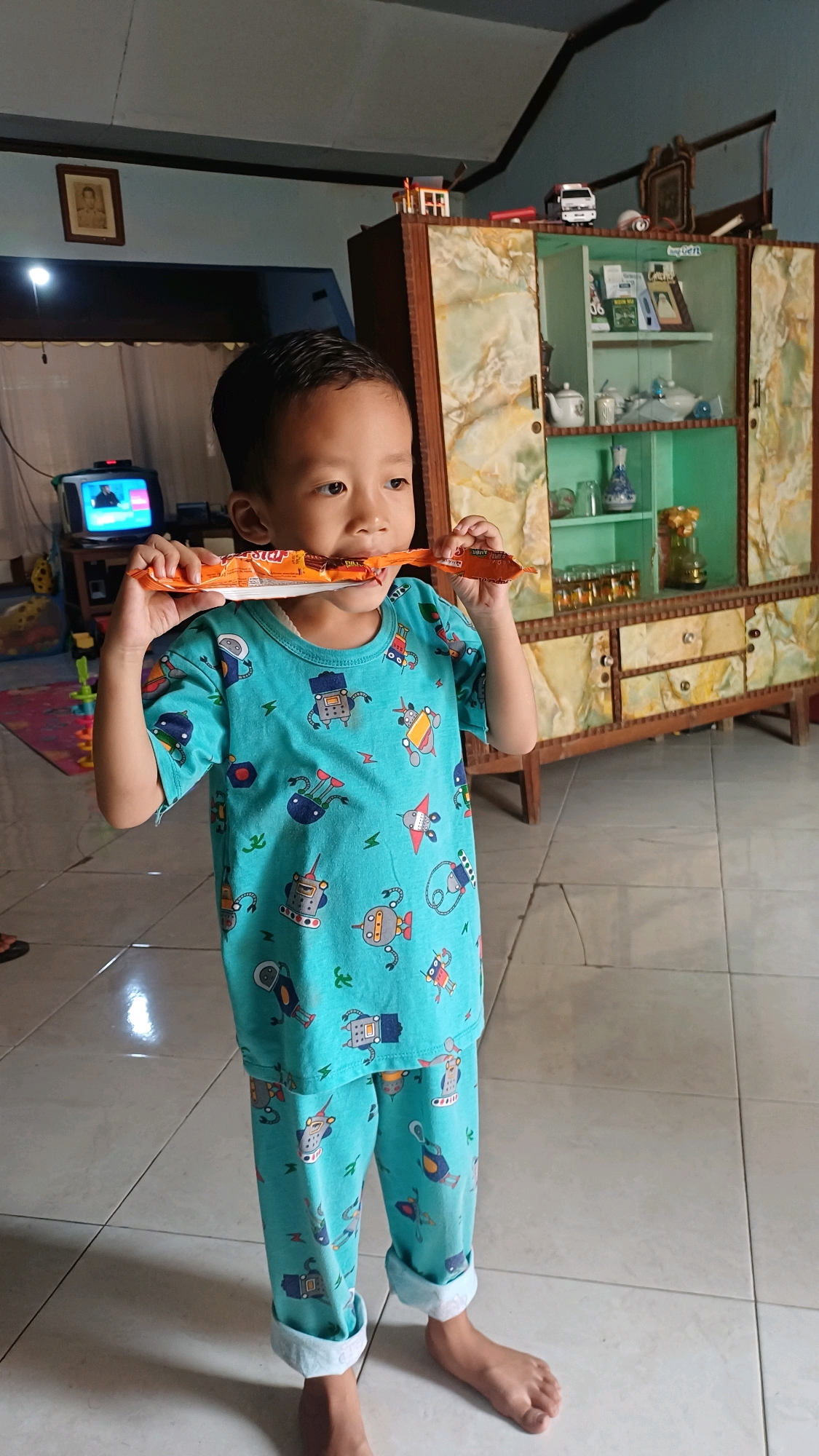 A young boy in teal pajamas with a robot pattern stands in a room, biting into a long, orange snack. The room appears to be a living area, with a television set on a stand to the left and a large, ornate wooden cabinet filled with decorative items to the right. The cabinet has glass doors revealing shelves stocked with various objects, including teacups and bottles. The floor is tiled in a light color. The boy is looking towards the right of the frame, and his hands are holding the snack up to his mouth. The overall impression is of a casual, domestic scene. The location is identified as Wonokerso, Indonesia.