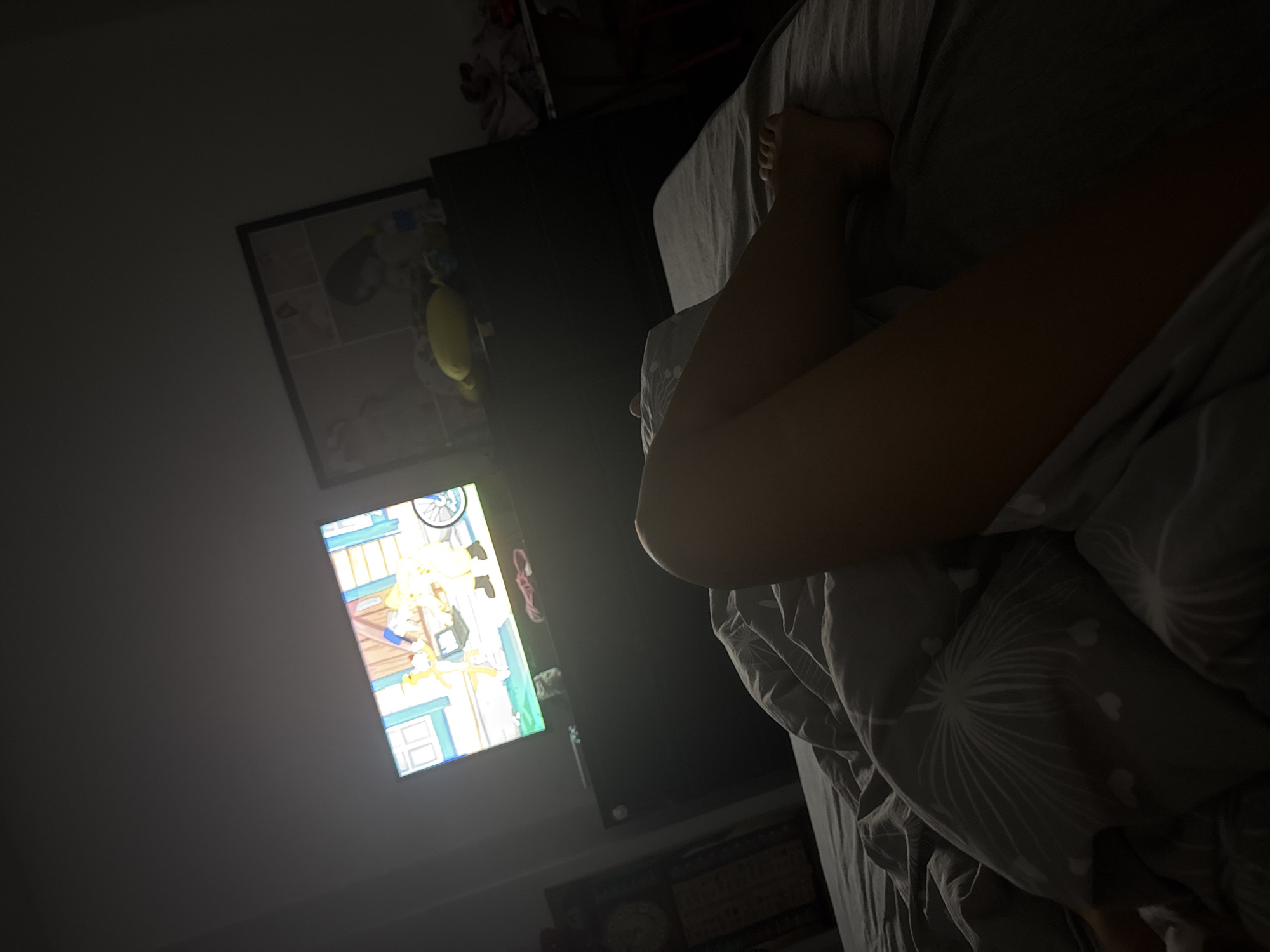 In a dimly lit bedroom, a person's legs, covered by a gray patterned duvet, are the focal point. A television screen in the background displays a cartoon with yellow characters, casting a faint glow. To the right of the TV, a framed collage of images, possibly photographs, rests on a dark dresser. A yellow plush toy is also visible on the dresser. The room appears to be a residential setting in Chișinău, Moldova, though no specific landmarks are identifiable to confirm this location. The overall impression is one of a quiet, relaxed evening spent in bed watching television.