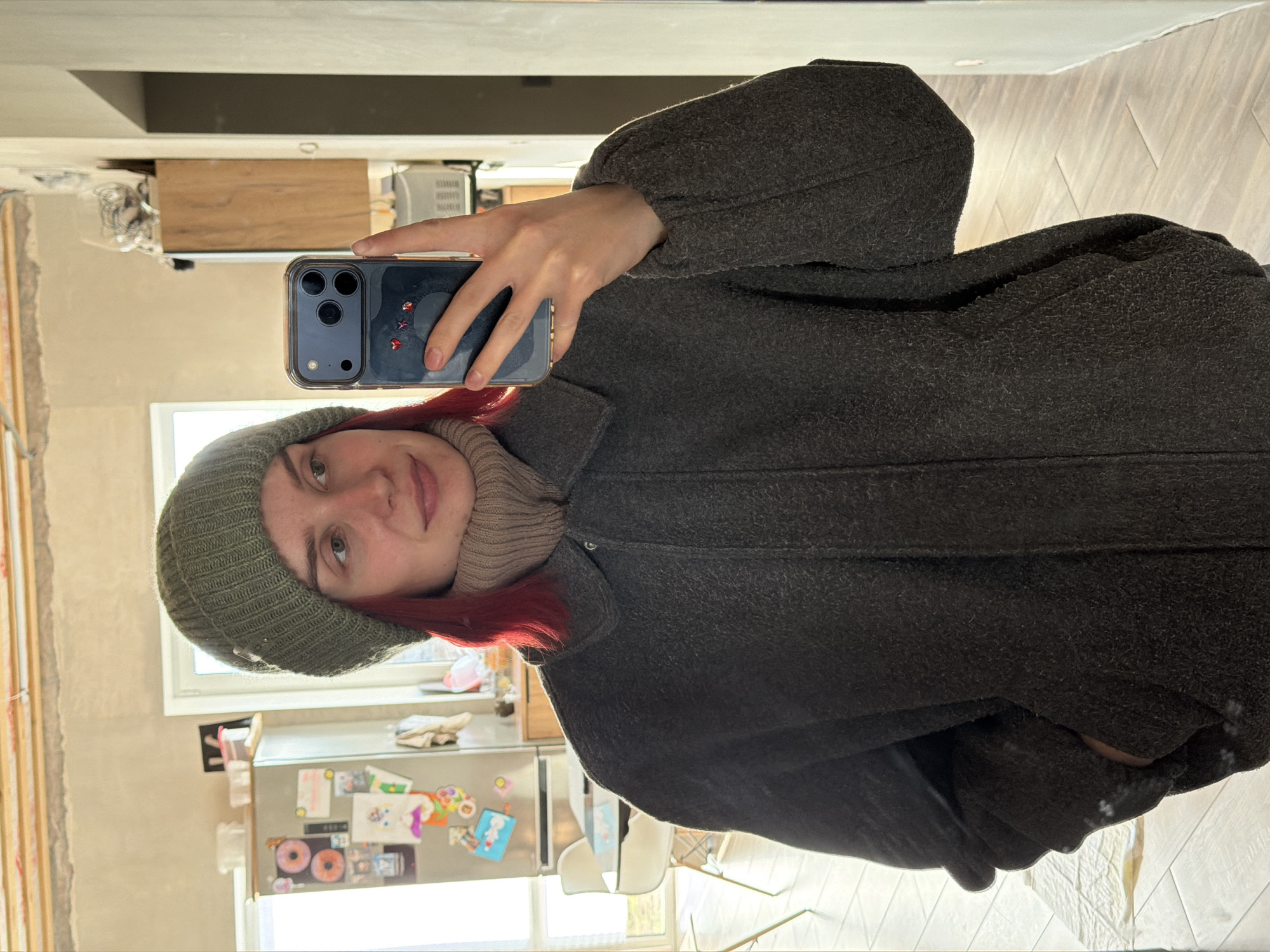 A young woman with red hair wearing a green beanie, brown turtleneck sweater, and charcoal wool coat is taking a mirror selfie. The phone she is using has a blue case with star decorations on the back. The environment appears to be a room undergoing renovation, with exposed wall framework visible behind her. A refrigerator covered in magnets and children's artwork stands against the wall. A table and white chairs are visible in front of a window. Based on the prompt stating the city of Chaikovsky, Russia, is relevant to the image, the location of the photograph is in this city.