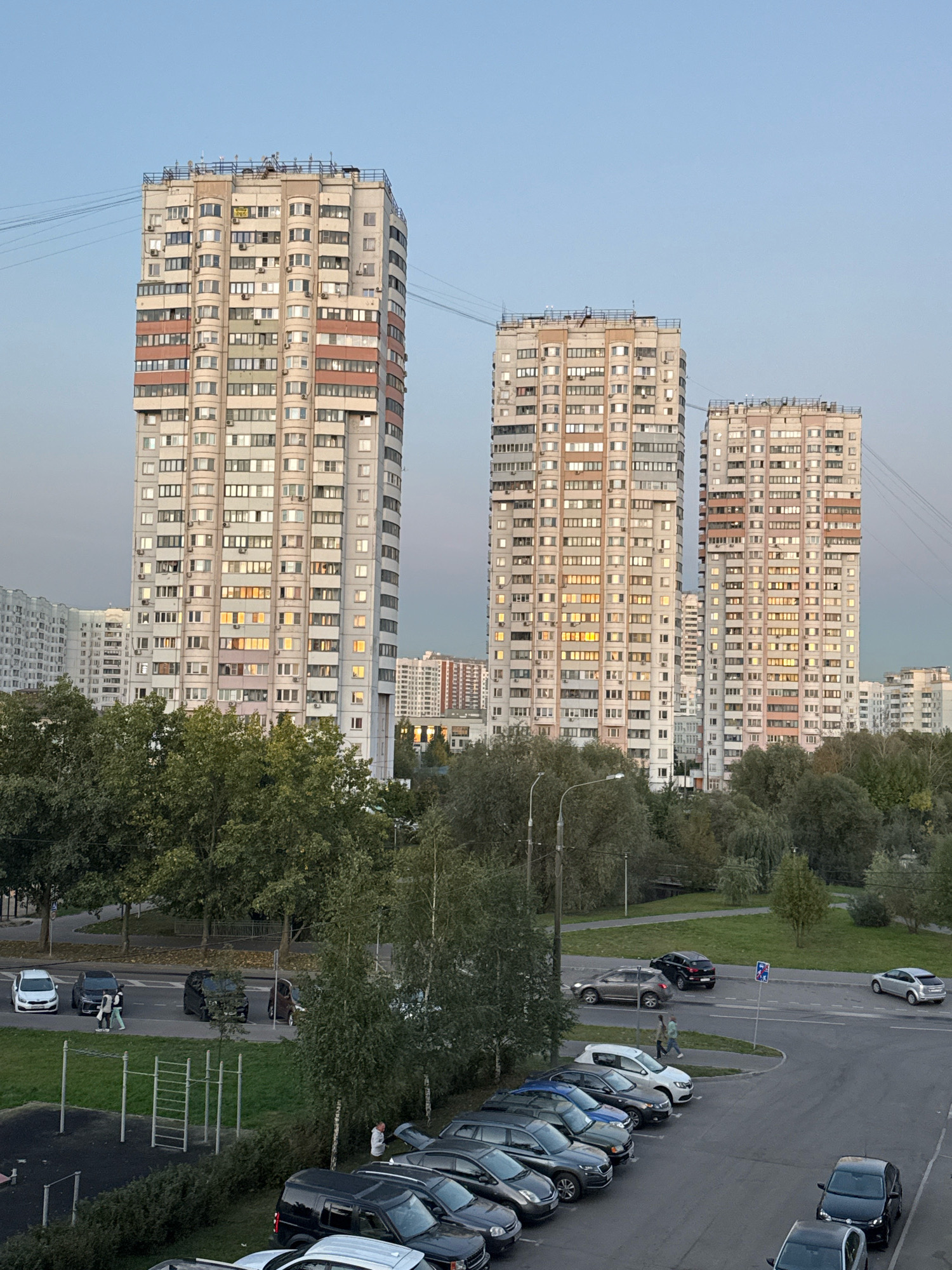 In Милицейский посёлок, Russia, the image depicts a view of several tall, multi-story apartment buildings against a pale blue sky. The buildings are rectangular in shape and have many windows, with some windows illuminated by interior lights, suggesting it is late afternoon or early evening. Several smaller apartment buildings and trees are visible in the middle ground. In the foreground and to the right, there is a parking lot filled with various cars, including sedans, SUVs, and smaller vehicles. Some of the car trunks are open. A street with passing cars and pedestrians is visible to the left of the parking lot, alongside a green grassy area with a children's playground structure. The overall scene conveys a typical urban residential environment in Russia.