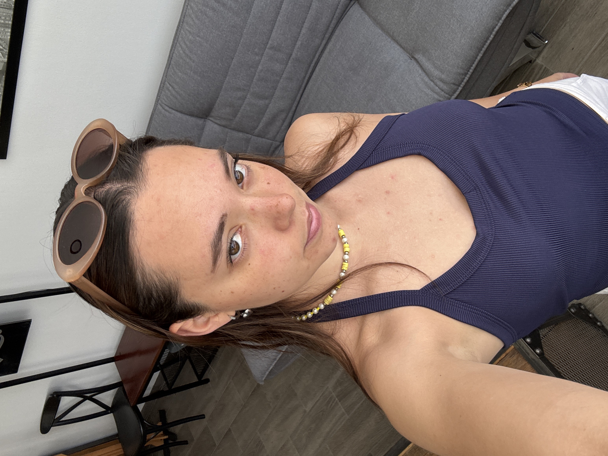A woman with brown hair and tan skin is taking a selfie in a room with neutral tones. She is wearing a navy blue tank top and a yellow beaded necklace. Sunglasses are perched on her head. The background includes a gray upholstered couch, a dark wood and metal side table, and a black metal chair. The floor is tiled in a light gray pattern. There is insufficient information to determine if the location is in San Bernardino Tlaxcalancingo, Mexico.