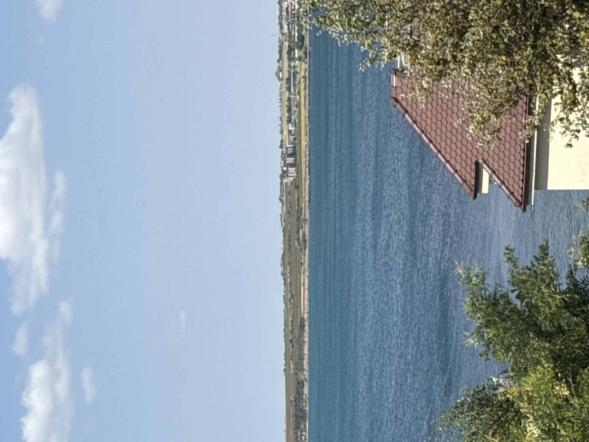 The image is a landscape view of a body of water with a distant shoreline. In the foreground, the roof of a building and some trees are visible. The sky above is blue with scattered white clouds. The distant shoreline appears to be a mix of vegetation and buildings. This scene is located in the city of Pomorie, Bulgaria.