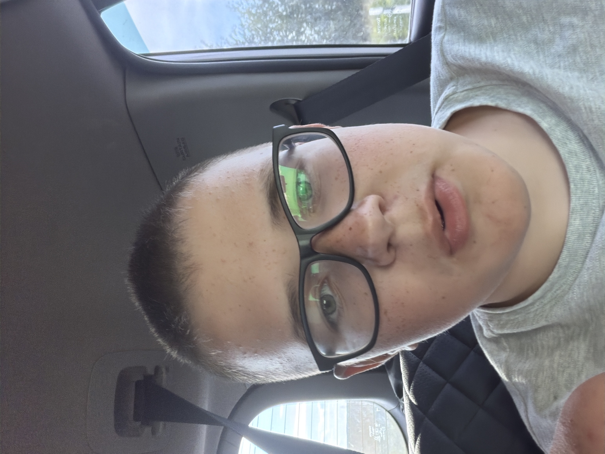 This is a close-up, selfie-style photograph of a young person wearing black-rimmed glasses and a grey t-shirt. The person has short, light brown hair and freckles on their face. They are looking directly at the camera with their mouth slightly open. The background suggests they are in a car, with a seatbelt visible behind them and a window on the right side showing trees and a cloudy sky. The interior of the car appears to be grey. The image does not contain information that would allow for specific localization to the city of Новое Маврино, Russia.
