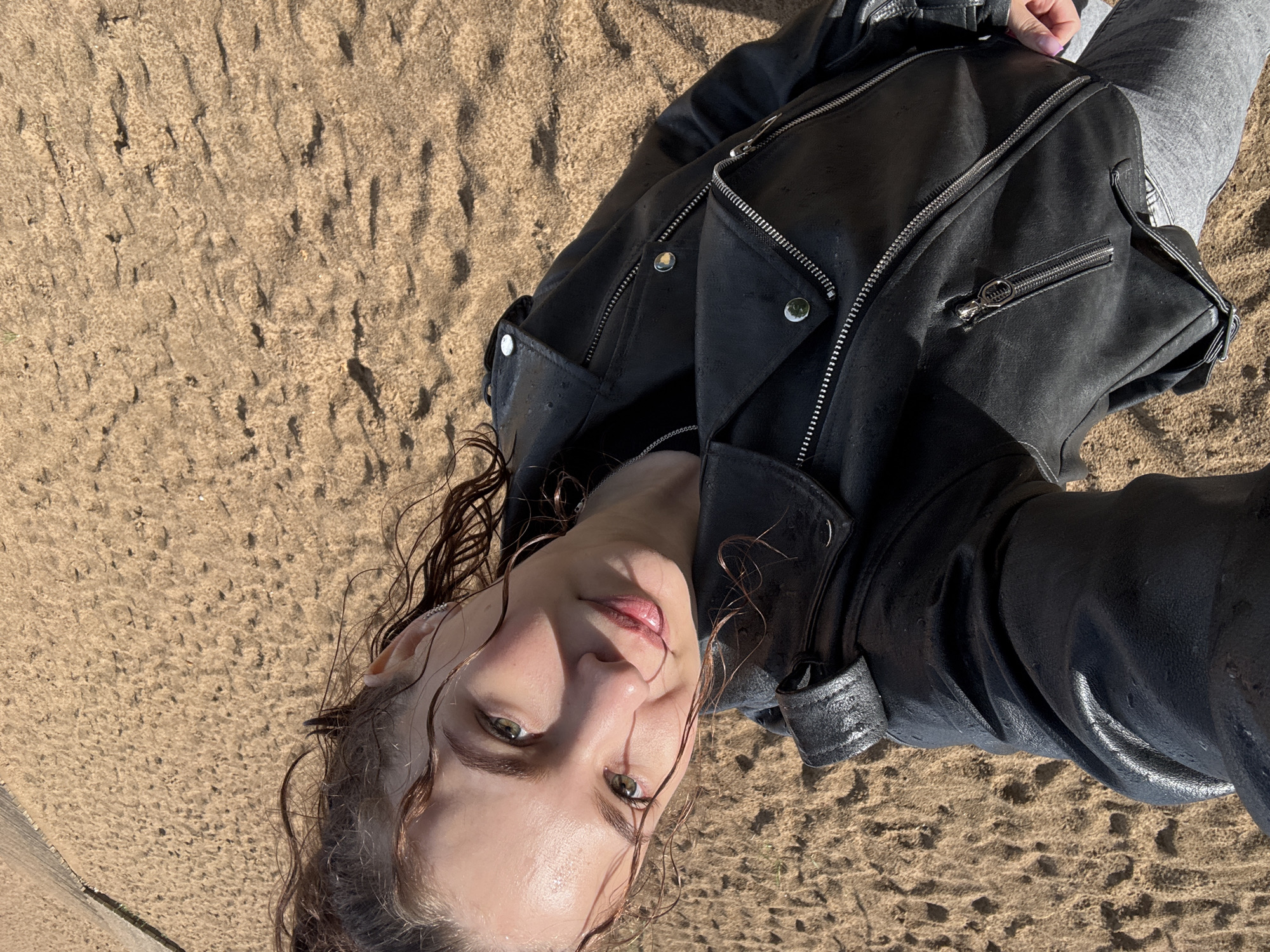 The image captures a person lying on their back on textured sand, looking directly up at the camera. The individual, a young adult, has light skin, wet, wavy brown hair, and appears to have green or hazel eyes. Their hair is visibly damp, with strands clinging to their face and forehead. A small silver ear cuff is visible on their left ear.
They are dressed in a black leather biker-style jacket adorned with prominent silver zippers and snaps. Below the jacket, gray casual pants or sweatpants are visible. A hand with light-colored nail polish is also seen.
The setting is an outdoor sandy environment, characterized by fine sand with undulating ripples, possibly formed by wind or water, suggesting a beach, riverbank, or sand dune. The scene is brightly illuminated by direct sunlight, casting distinct shadows and highlighting the various textures of the sand and the leather jacket. The bright lighting indicates it is daytime.
No visible text is present in the image. The overall impression is one of a relaxed pose in a natural, sunlit environment. Given the location context of Chaikovsky, Russia, this sandy area could be along the Kama River or a nearby body of water.