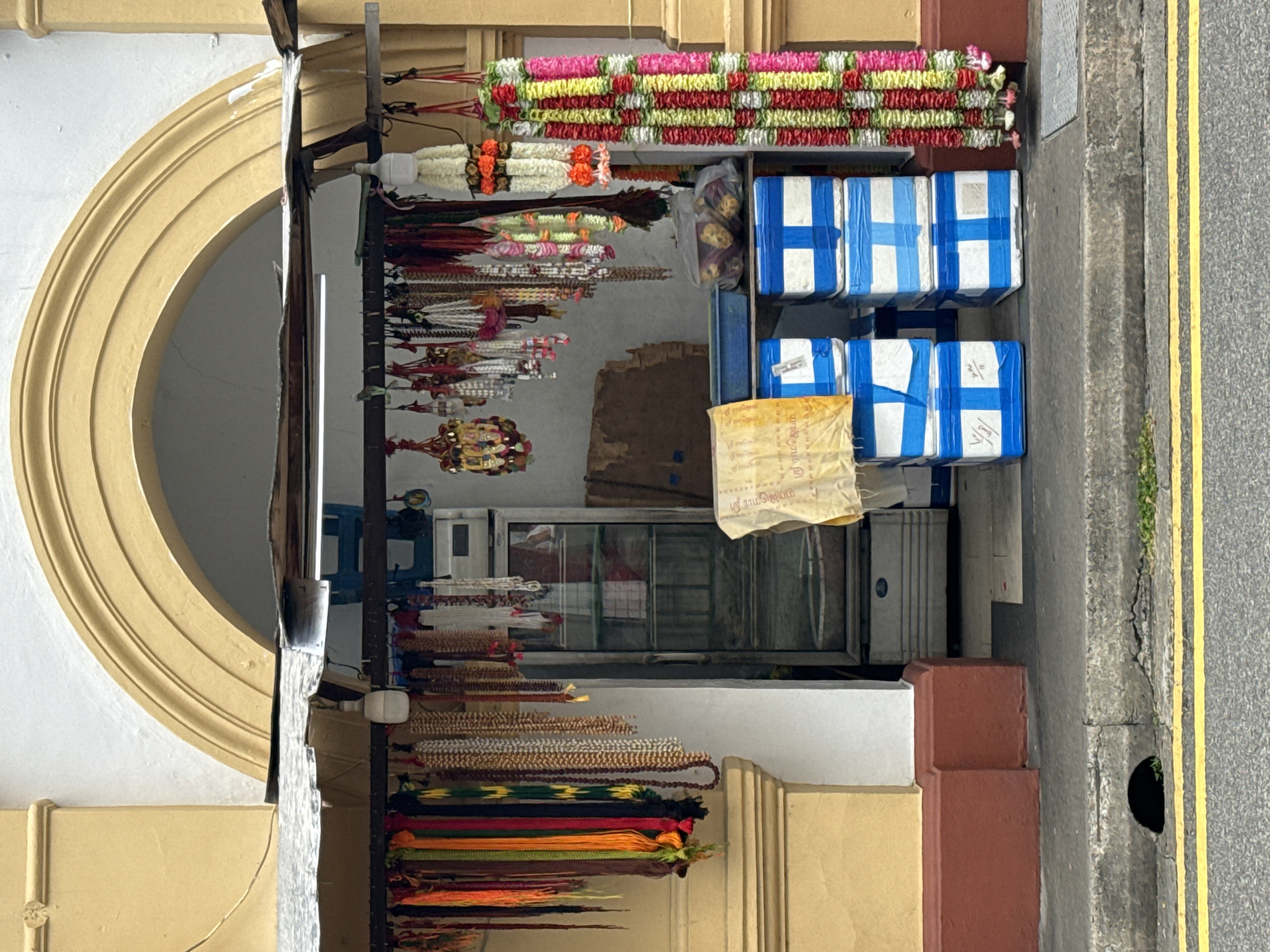 The image shows a street-level view of a small shop in Singapore, likely selling religious items. The shop is located under a yellow arched facade, with a corrugated metal awning extending over the entrance. Numerous garlands and beaded necklaces in vibrant colors hang from a rack above the shop's interior. Inside the shop, a glass-fronted refrigerator is visible, along with stacks of blue and white boxes that appear to be sealed with tape. A yellow cloth with Tamil writing is draped over some of the boxes. The shop is situated on a sidewalk next to a street with yellow double lines, suggesting it is a public thoroughfare. The overall scene depicts a typical urban setting in Singapore, characterized by its blend of traditional and modern elements.