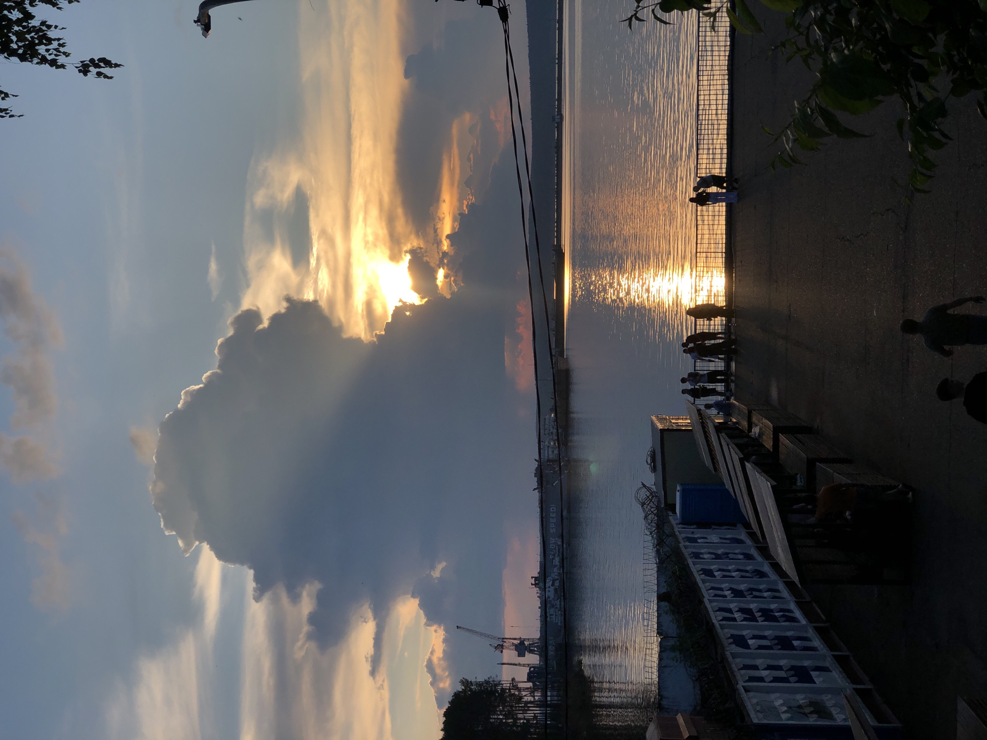 The image depicts a sunset over the Kama River in Chaikovsky, Russia. The sky is filled with dramatic clouds, with the sun peeking through, casting a golden glow on the water. Silhouetted figures of people can be seen on the riverbank, enjoying the view. In the background, industrial cranes and buildings are visible, hinting at the city's port and industrial activity. A fence with barbed wire runs along the riverfront, separating the walkway from the water. Several market stalls are set up along the walkway, some with awnings providing shade. The overall scene captures a serene moment as day transitions into evening in a Russian industrial city.