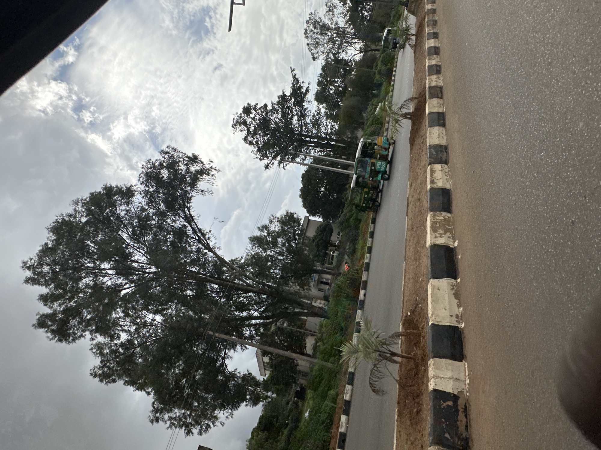 This media file depicts a street scene in Bar Jirgi Summa, Nigeria. The image is oriented vertically, with the road and its adjacent curb occupying the right side of the frame. The road surface is paved asphalt, speckled with small stones. Along the edge of the road is a curb with alternating black and white rectangular blocks, some of which appear weathered and chipped.
On the left side of the road, a lush green landscape unfolds, dominated by several large, mature trees with dense foliage. The trees are tall and their branches spread widely, creating a significant canopy. Interspersed among the trees are some buildings, appearing to be residential or commercial structures.
Several auto-rickshaws, commonly known as "Keke Napep" or "Marwa," are parked or moving along the road. Two are clearly visible in the middle ground, painted in a green and white color scheme. One has visible text on its side, including "PTA 10255". Another similar vehicle is further down the road, partially obscured by trees. The presence of these vehicles suggests this is a populated area with local transportation.
The sky above is overcast with a mix of white and gray clouds, indicating a potentially cloudy or a recent rainy day. The lighting suggests it is daytime, likely late morning or afternoon, given the diffused sunlight. A faint silhouette of a structure, possibly a traffic light or utility pole, is visible in the upper portion of the frame. The overall mood is quiet and observational, capturing a typical street view in a Nigerian locale.