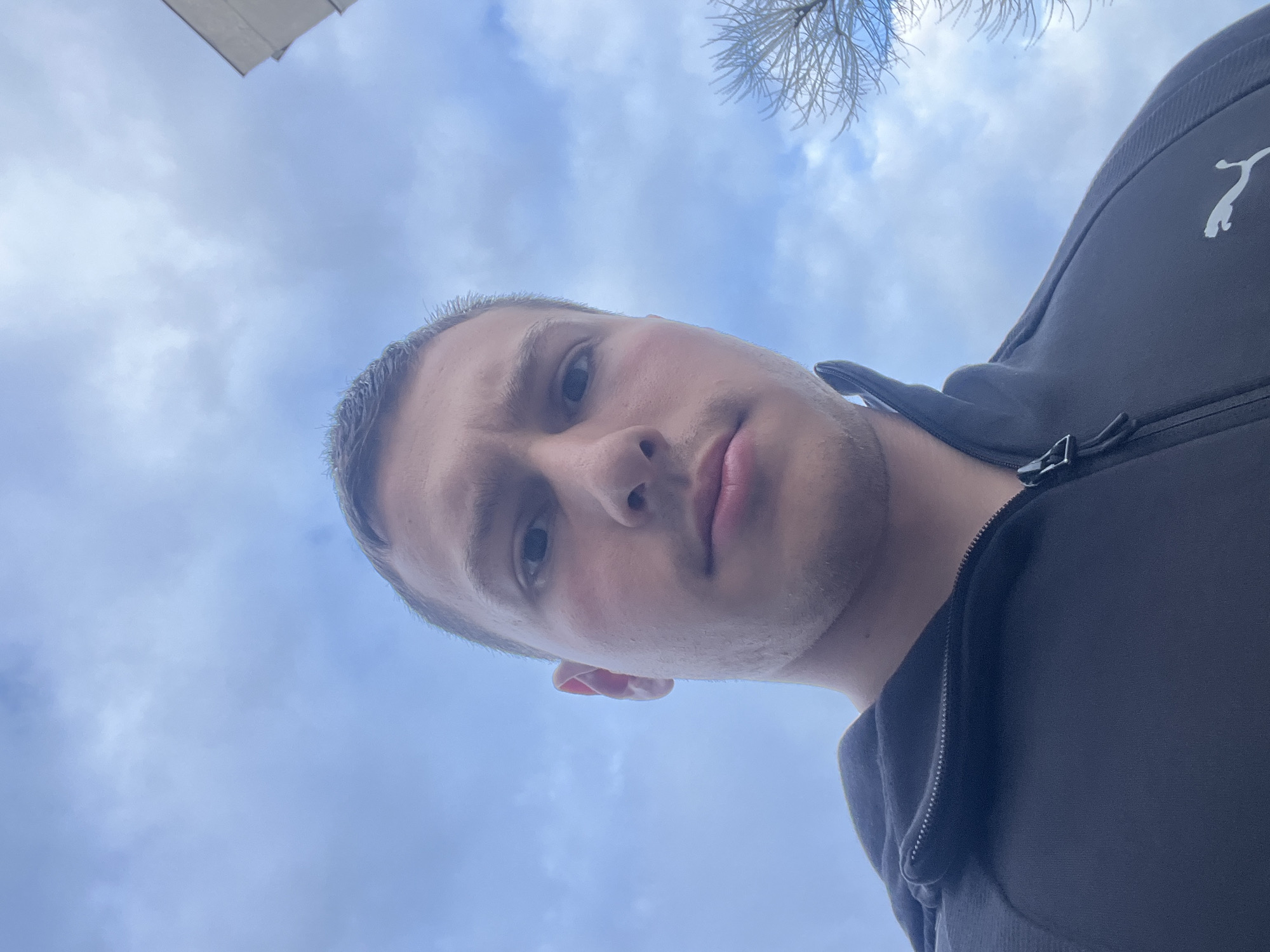 A low-angle selfie of a young man with short hair and fair skin. He is wearing a black zip-up Puma jacket. The background consists of a cloudy sky. It is not possible to determine the location in Burgas, Bulgaria, from the image alone.