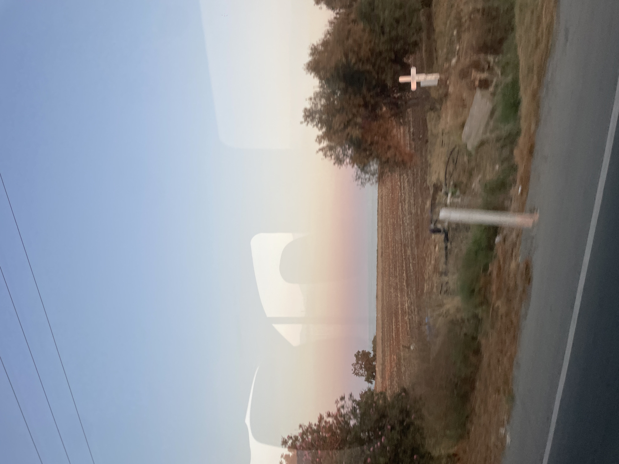 This image captures a rural landscape, likely near Timi Community, Cyprus, during what appears to be sunset. The sky transitions from a clear blue at the top to soft hues of pink and orange near the horizon, suggesting the golden hour. In the foreground, a paved road runs horizontally along the bottom of the frame, with a grassy verge bordering it.
The middle ground features a cultivated field, its texture indicating recent plowing or tilling. To the left, a bushy tree with pink flowers stands out against the dry, brown vegetation. Further back, a lone, leafless tree is visible in the distance. On the right side of the frame, a cluster of dense trees forms a backdrop for a white cross, positioned on a slightly elevated patch of ground. The cross is simple, with a horizontal bar meeting a vertical one, and a small plaque or inscription is attached to its lower part.
Reflections are visible on the surface from which the photo was taken, obscuring parts of the scene and adding a layer of abstraction. These reflections create abstract shapes and hints of interior elements. The overall atmosphere is serene, with a touch of melancholy conveyed by the presence of the cross and the muted tones of the landscape. There are no people clearly depicted in the scene.