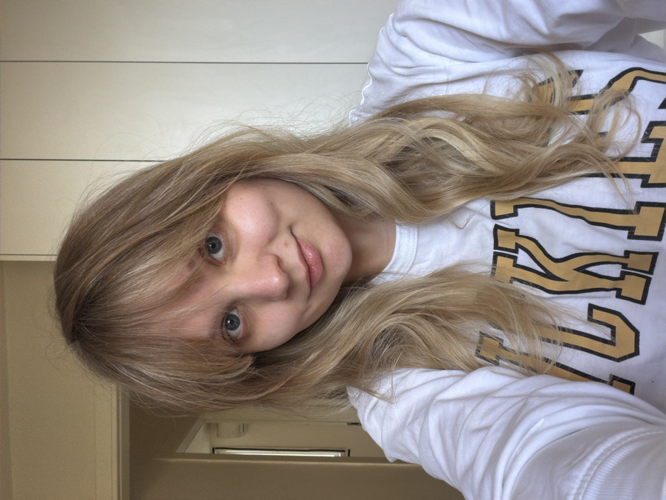 An indoor scene in Tel-Aviv, Israel, depicts a light-skinned female individual. She possesses long, wavy blonde hair with bangs, blue eyes, and a slight smile while looking directly at the camera. She is wearing a white long-sleeved shirt with "LAKERS" printed in gold and black block letters across the chest. The background on the left reveals a partial view of an interior doorway or hallway with a faint, light-colored object visible high on the far wall. The background on the right consists of a plain, light-colored wall or paneling with distinct vertical seams.