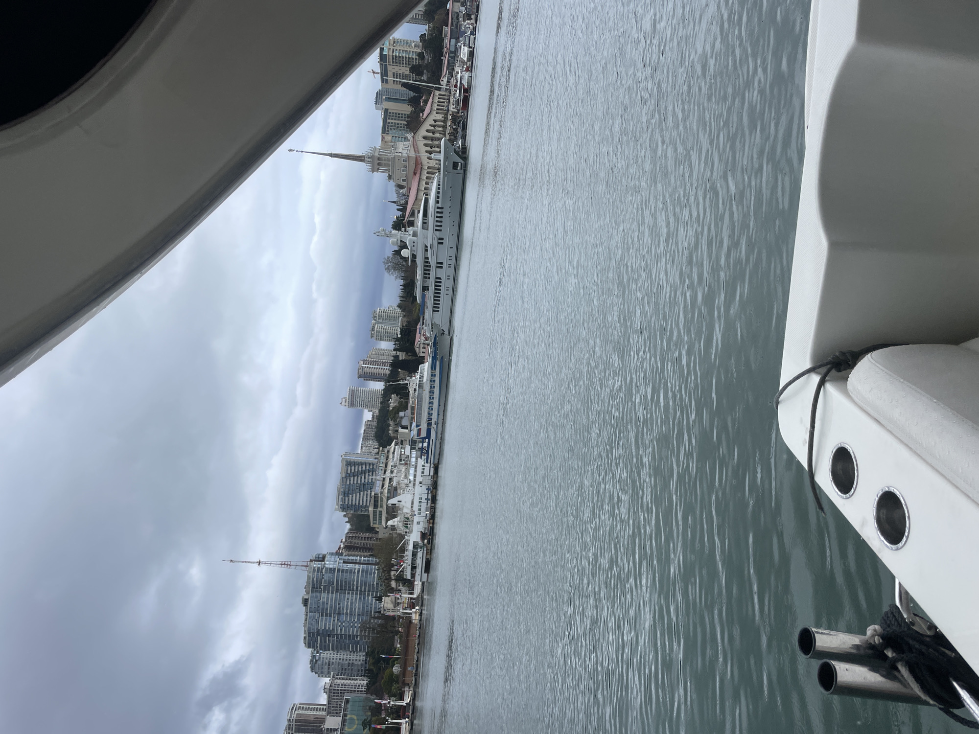 The image shows a view from a boat towards the city of Sochi, Russia. The sea is in the foreground, with the coastline featuring buildings, including tall residential complexes and the iconic Sochi Sea Port building recognizable by its spire. Several ships are docked at the port. The sky is overcast. Parts of the boat are visible in the foreground of the image.