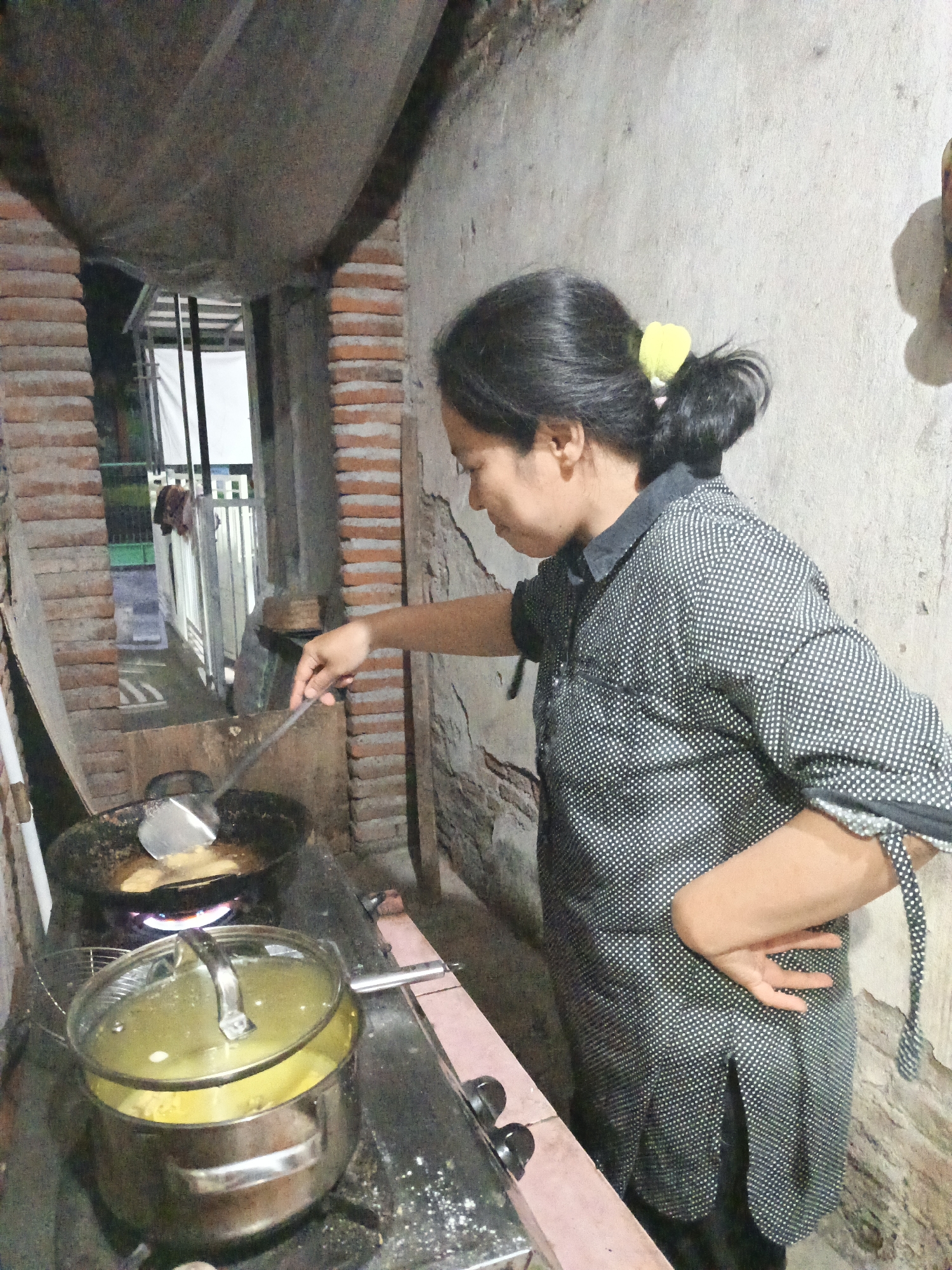 In Kepulungan, Indonesia, a woman with dark hair tied in a bun with a yellow scrunchie is cooking in a rustic outdoor kitchen. She is wearing a black and white polka dot button-down shirt and is standing in front of a gas stove. On the stove, a wok with a spatula is being used to fry food, and a stainless steel pot with a glass lid, containing yellow liquid, is also present. The kitchen walls are made of rough, aged concrete and brick, and the ceiling appears to be constructed from rough-hewn wood and concrete. In the background, through an opening, a glimpse of what appears to be an outdoor area or alleyway can be seen, with metal bars and some laundry hanging. The overall environment suggests a simple, perhaps traditional, living space.
