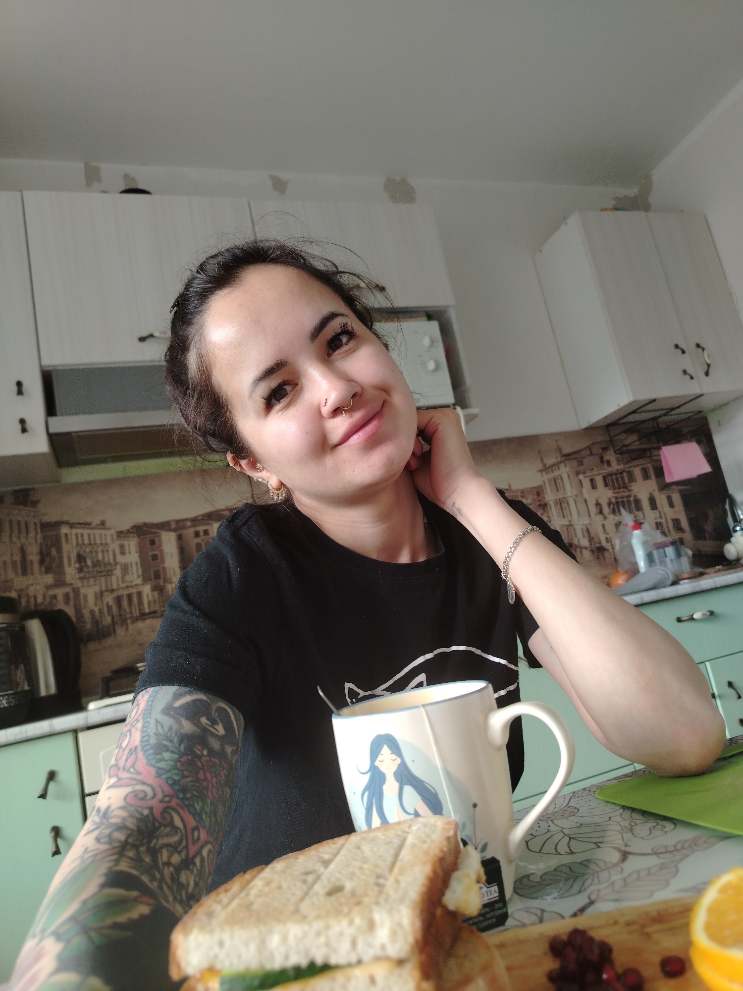 A young woman with dark hair pulled back, wearing a black t-shirt with a white design and a silver bracelet, is taking a selfie in what appears to be a kitchen. She has a nose ring and several earrings, and a large tattoo on her left arm and shoulder. She is smiling and resting her chin on her hand. In front of her, on a patterned tablecloth, is a sandwich, a mug with a blue design, some red berries, and a slice of orange. The background shows white kitchen cabinets, a microwave, and a tiled backsplash with a cityscape pattern. The lighting suggests it is daytime. The location is not specified in the image or provided context.