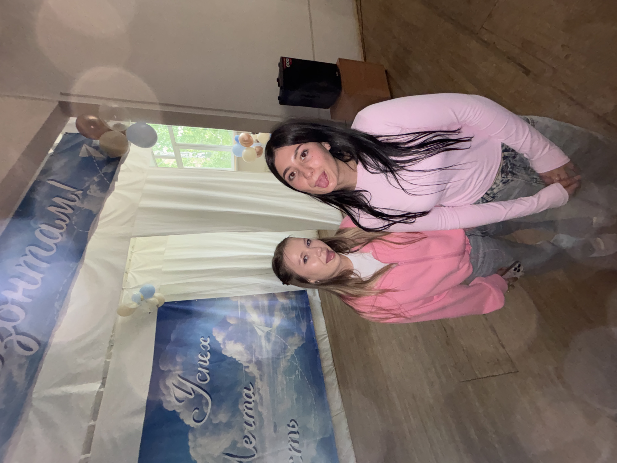 The image depicts two young women standing in what appears to be an indoor event space, possibly a school assembly hall or community center, located in Chaikovsky, Russia. The backdrop includes a large banner with text in Cyrillic, suggesting a celebratory or motivational theme, with imagery of clouds and a sky. Balloons are visible, adding to the festive atmosphere. The women are casually dressed in pink tops and jeans. One woman has blonde hair and is wearing a pink zip-up jacket, while the other has long, dark hair and is wearing a pink long-sleeved shirt. Both women are sticking out their tongues towards the camera, indicating a playful mood. In the background, a black speaker sits on a wooden box. The floor is made of wooden planks. The overall impression is of a casual gathering or celebration in a public indoor venue.