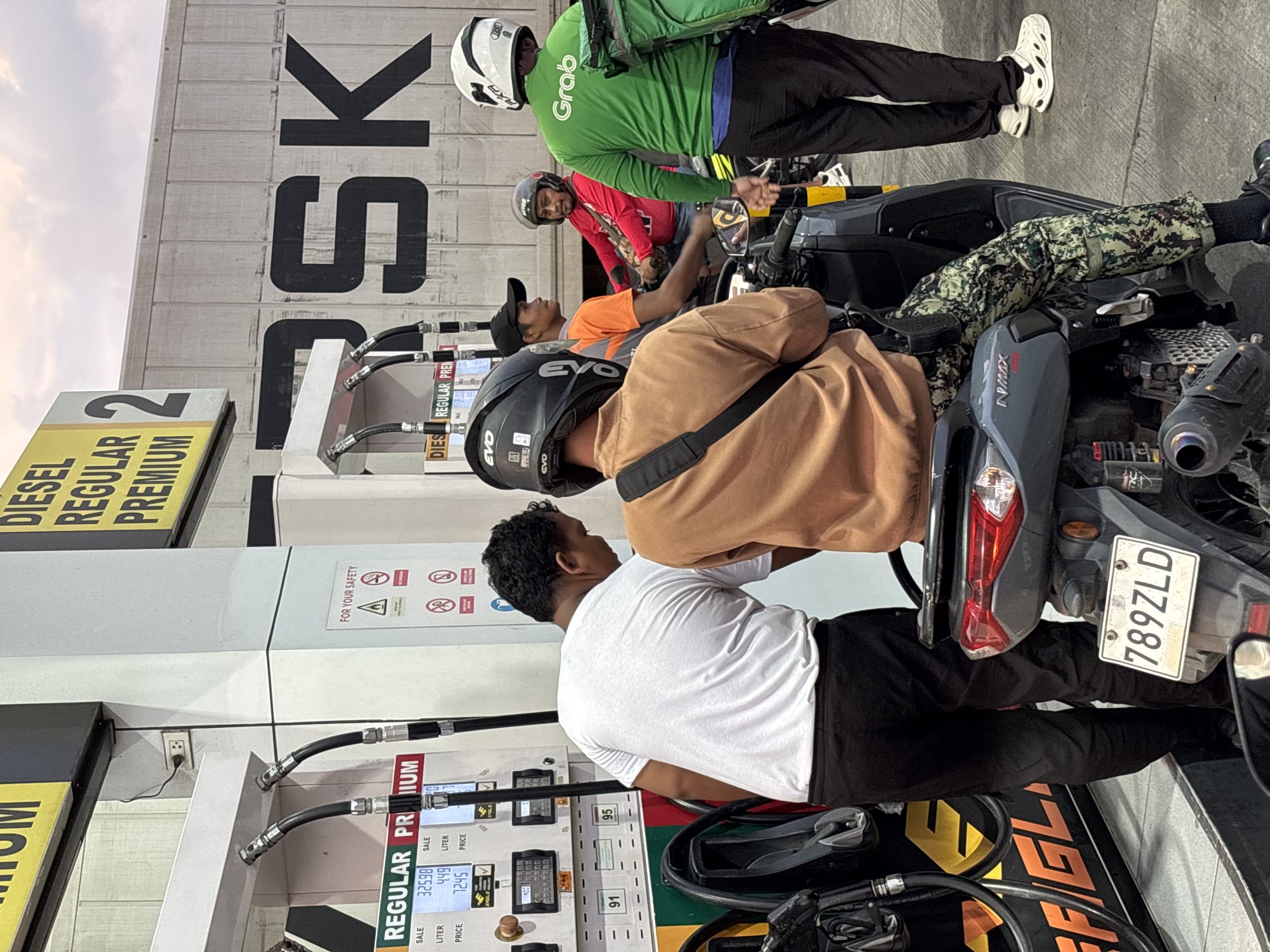 The image depicts a scene at a gas station in Davao City, Philippines. In the foreground, two individuals are present. One, wearing a tan long-sleeved shirt and camouflage pants, sits on a motorcycle facing away from the camera. The other person, wearing a white t-shirt and black pants, stands next to the motorcycle, appearing to interact with a gas pump.
The gas pump displays prices and volumes for different fuel types, with "REGULAR" and "PREMIUM" visible. Several gas hoses are connected to the pump. Behind the people, a large concrete wall with black lettering that reads "RSK" is visible. Further in the background, a person in a green Grab uniform is visible, also with a motorcycle.
The overall environment suggests an active fuel station with multiple customers and motorcycles. The sky above is overcast, indicating a potentially cloudy or evening time. The presence of "Grab" suggests a ride-sharing service is operating in the area. The license plate on the motorcycle in the foreground reads "789ZLD".