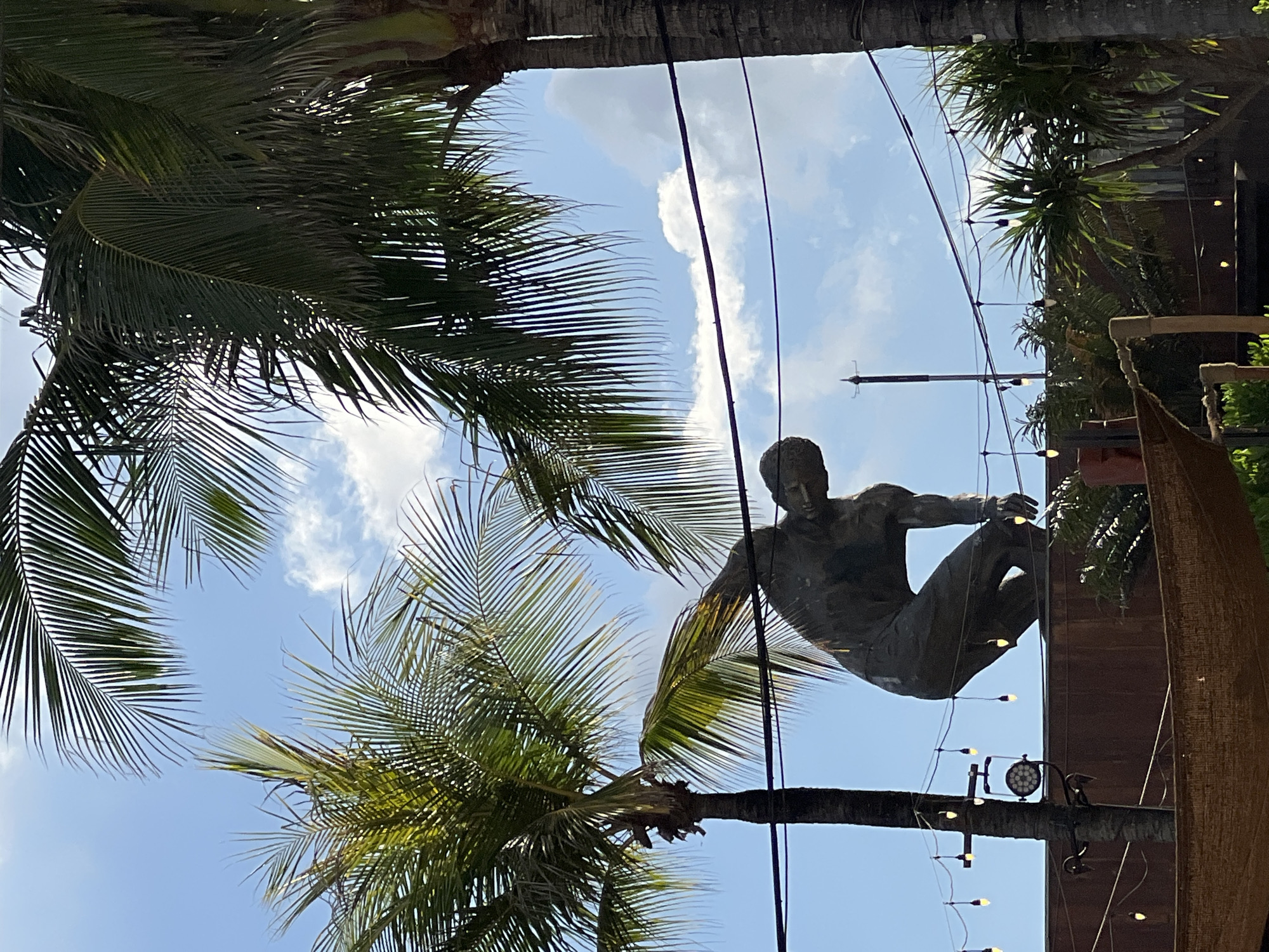 In the city of North Kuta, Indonesia, a large bronze statue of a man in a crouched, contemplative pose stands on a dark brown platform under a bright blue sky with scattered clouds. The statue is positioned between two prominent palm trees, their fronds obscuring parts of the sky. Overhead, strung with small, incandescent bulbs, are decorative lights that appear to be strung between poles or structures. The overall scene suggests an outdoor venue, possibly a park or a resort, with a focus on tropical aesthetics and public art.
