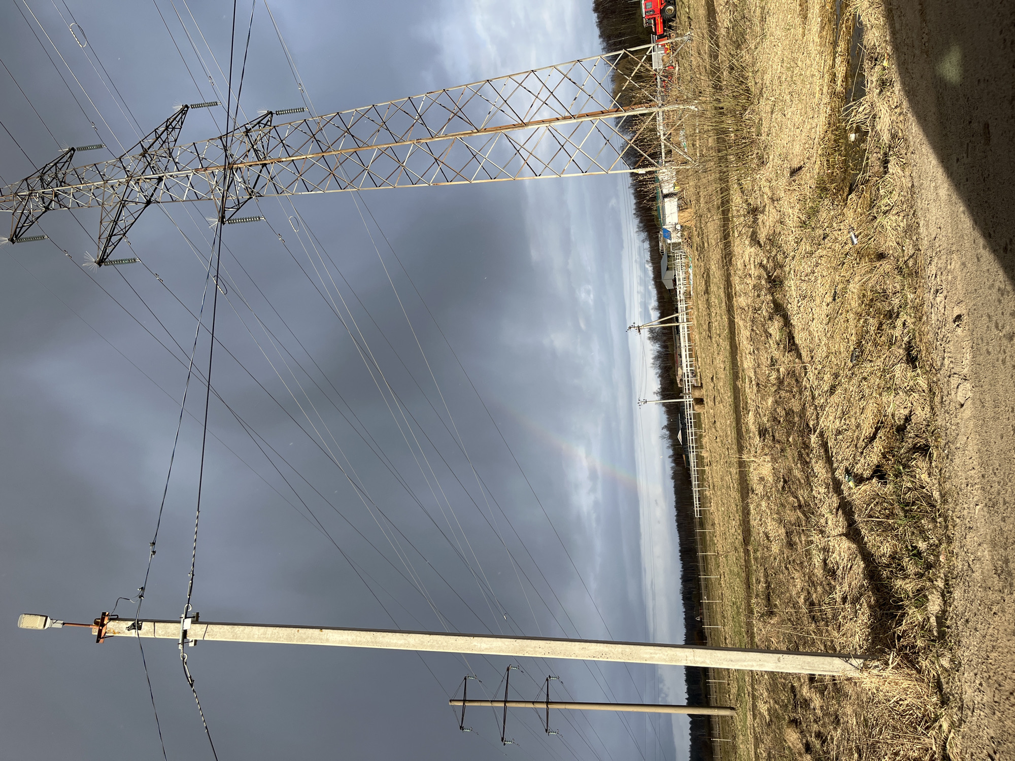 The image depicts a rural landscape in Razdol'e, Russia, dominated by power lines and utility poles against a cloudy sky. A large, metal lattice transmission tower stands prominently in the background. Closer to the foreground are concrete utility poles with insulators and electrical wiring. The ground is covered in dry, brown grass and vegetation. In the mid-ground, a faint rainbow arc appears in the sky. In the far background are some small structures, a line of trees, and possibly a red vehicle. The overall scene is typical of an agricultural or industrial area, with infrastructure serving the region.