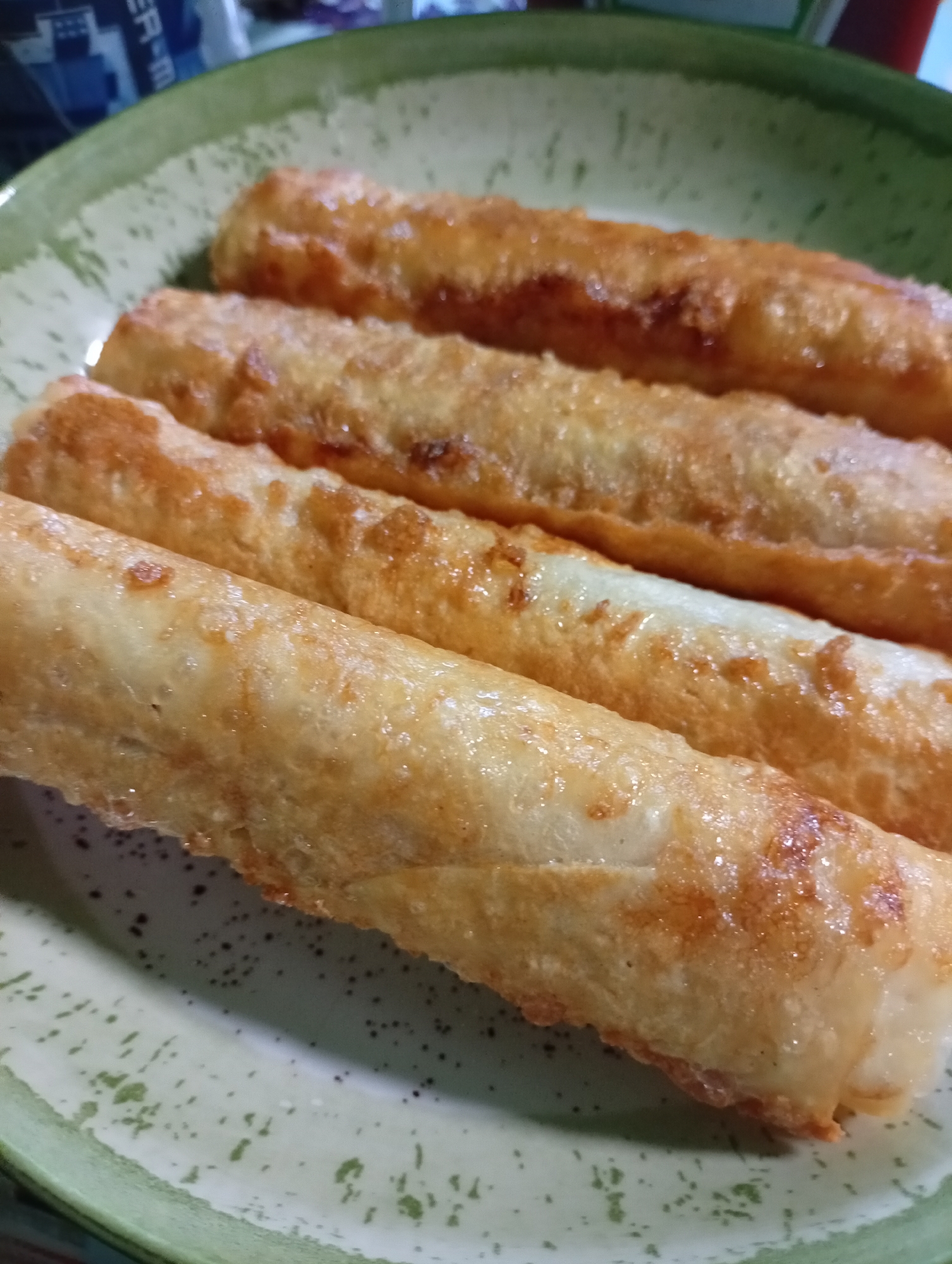 A close-up, slightly angled shot shows four golden-brown, cylindrical fried pastries arranged horizontally on a light green speckled plate. The pastries appear to be spring rolls or similar fried dough items, with some visible oil sheen on their surfaces. The plate has a textured rim with a darker green border. In the upper left corner, out of focus, are glimpses of packaging, including what appears to be blue and white text. There are no people or animals visible in the image. The scene is indoors and appears to be a domestic setting. There is no information in the image to suggest the location is Pavlovsky Posad, Russia.