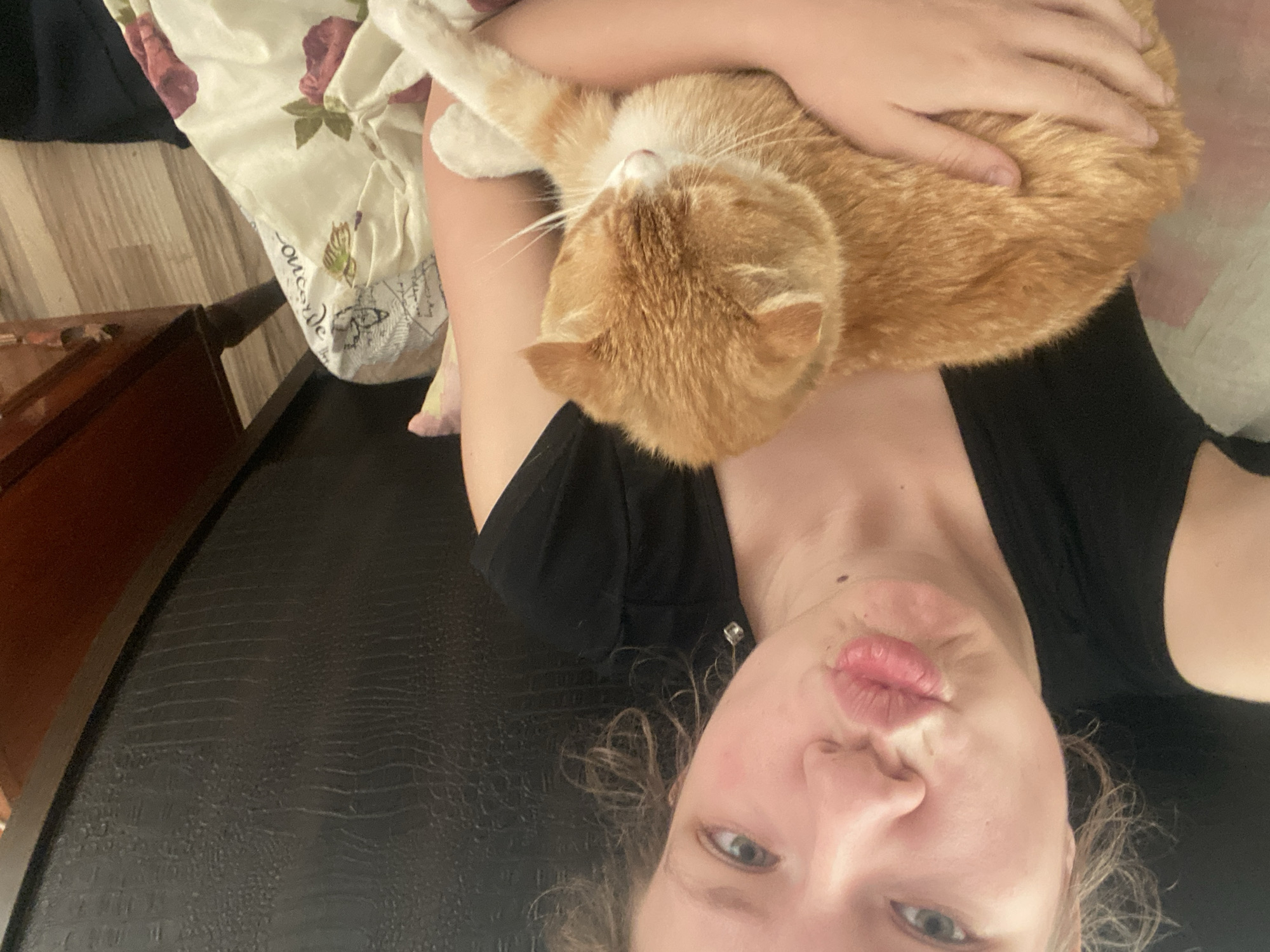 A person is lying on a surface, holding an orange tabby cat. The person is wearing a black shirt. The cat is resting its head and body on the person's arm and chest. The background includes a floral-patterned blanket/sheet and a wooden piece of furniture. The surface the person is lying on has a dark, patterned texture. There is no information to indicate this scene is located in Pakhynia, Ukraine.