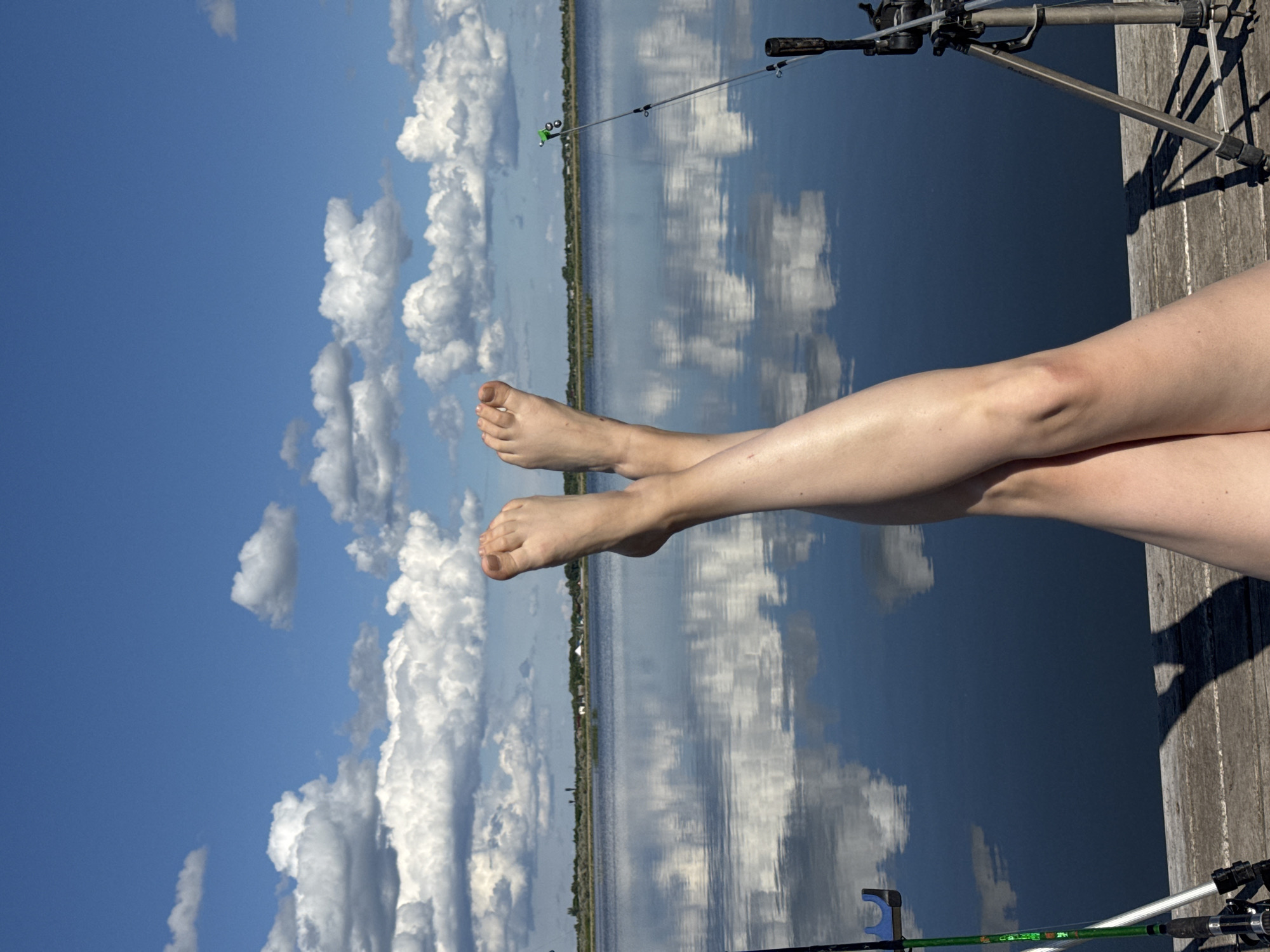 A perspective shot taken in the Unknown, Unknown shows a pair of human legs extending from the bottom right corner of the frame, crossed at the knees, with the feet pointing towards the upper left. The legs appear to be resting on a wooden structure, possibly a dock or pier. In the background, there is a body of water, likely a lake or river, reflecting a clear blue sky with puffy white clouds. A fishing rod setup, including a tripod, is visible in the upper right corner. Another fishing rod is visible at the bottom. A treeline is visible on the horizon.