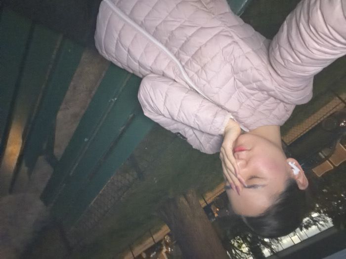 A young woman is seated on a dark green slatted park bench in an outdoor setting during twilight or nighttime, characteristic of a public green space in Paris, France. She wears a light pink quilted jacket with a white zipper and has a white wireless earbud in her left ear. Her right hand is raised to her face, with fingers spread across her forehead and right eye, while her left eye is closed. Her fingernails are painted a dark color. The background shows a parked dark-colored vehicle, a tree trunk, a wire fence, and distant architectural forms illuminated by artificial light, consistent with an urban park environment.