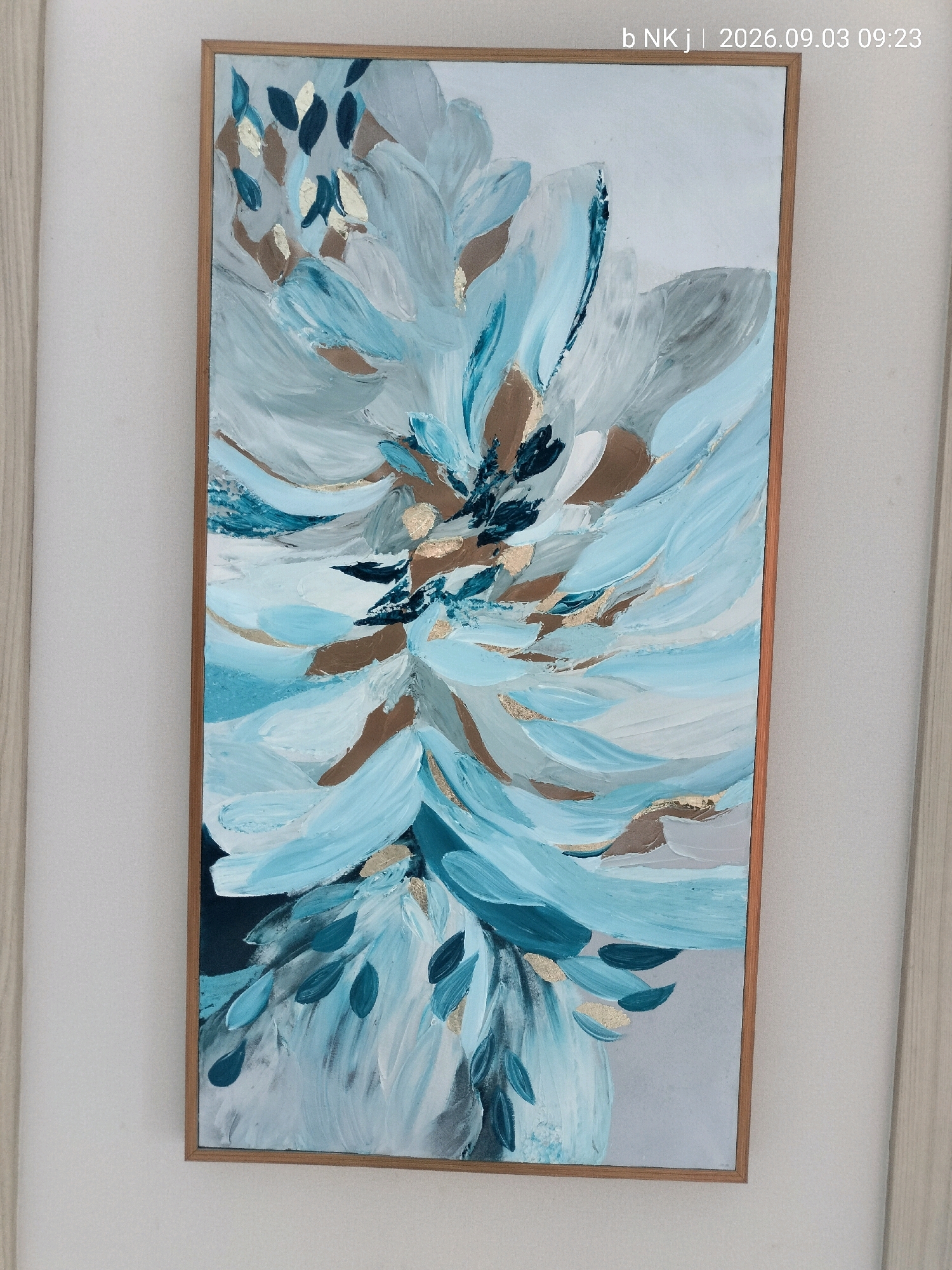 A framed abstract painting of a stylized flower or botanical element hangs on a white wall. The painting features a palette of blues, whites, grays, and metallic gold accents, with visible brushstrokes creating a textured effect. The frame is a light wood. The image also contains a timestamp in the upper right corner that reads "b NK j | 2026.09.03 09:23". There is no discernible location in the city of South Jakarta, Indonesia.