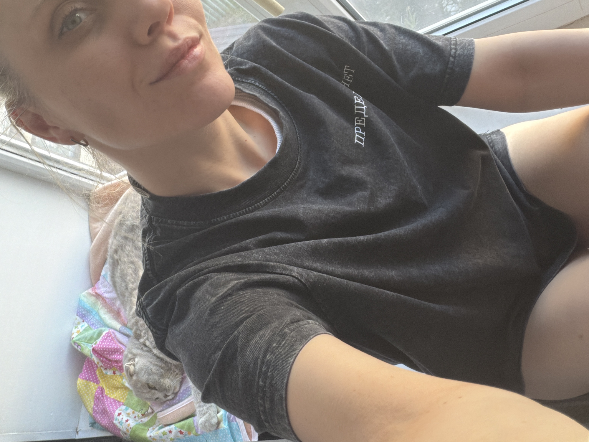 A woman is posing for a selfie indoors, with a grey cat resting on a colorful patchwork quilt next to her. The location appears to be near a window. The text on the shirt is not legible enough to determine the location in Chaikovsky, Russia. The woman is wearing a grey t-shirt.