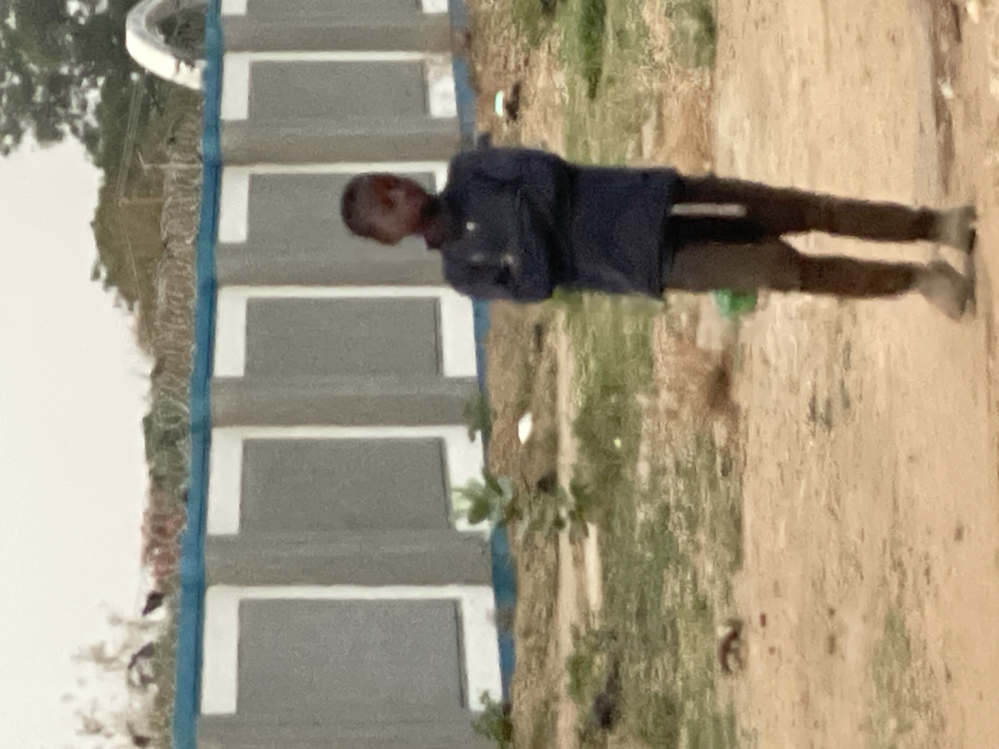 A person is standing outdoors in Hayin Banki, Nigeria, next to a building with a light-colored facade and blue trim. The person is wearing a dark blue shirt and dark pants. The ground is light brown and there is green vegetation present.