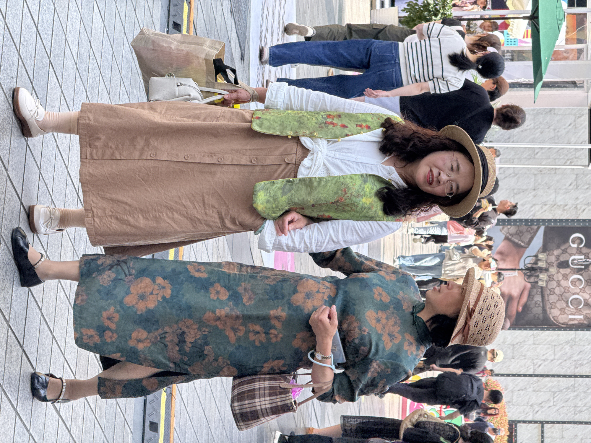 The image captures two women in the foreground on a bustling urban street, with numerous other people and buildings in the background. The scene is brightly lit, suggesting a sunny daytime.
The woman on the left, closer to the viewer, wears a white long-sleeved top tied at the waist, a long, light brown button-front skirt, and a green vest with a subtle floral or leafy pattern. She has dark, wavy hair, wears clear-rimmed glasses, and a light-colored straw hat with a dark band. She is smiling and appears relaxed, looking towards the right side of the frame. She carries a white shoulder bag and a large, light brown shopping bag. Her footwear consists of light-colored socks and white oxford-style shoes.
The woman on the right, slightly further back, is dressed in a long, dark teal or green dress adorned with a prominent pattern of large brown or orange flowers and leaves, possibly styled after a traditional Chinese cheongsam. She also wears a woven straw hat, distinguished by a pink ribbon. She has dark hair and is looking to her left. She carries a small plaid brown and white handbag on her arm and has a watch with a light blue band on her left wrist, while holding a smartphone in her right hand. Her shoes are black Mary Jane style with silver buckle straps.
The background is populated by many blurred figures, indicating a busy pedestrian area. A large advertisement is prominently displayed on a building or billboard, featuring an image of a handbag and the word "GUCCI" written vertically in large, dark letters. The street is paved with light-colored square tiles. The overall setting is a vibrant commercial or public space in an urban environment, likely in Yuzhong District, China.