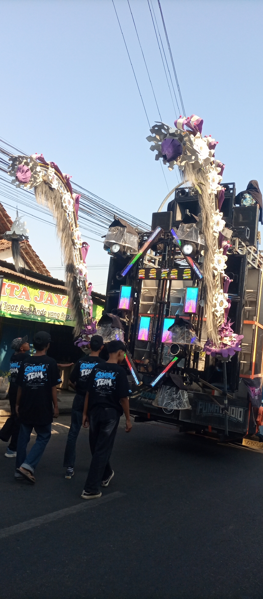 In Jekulo, Indonesia, a procession moves down a road under a clear sky. The central focus is a large, multi-tiered sound system mounted on a vehicle. This system is adorned with elaborate decorations, including large, fluffy plumes in shades of white and purple, interspersed with what appear to be fabric flowers and faux fur. The sound system itself features numerous speakers, lights, and digital displays that emit colored light. Several people, mostly young males wearing black t-shirts with the "SEMPAL TEAM" logo, are walking alongside or behind the sound system. They are dressed casually in jeans and sneakers. In the background, a building with a green awning and signage is visible. Overhead, a dense network of electrical wires crosses the sky. The road surface is asphalt, marked with a white line. The overall scene suggests a mobile entertainment event or parade.