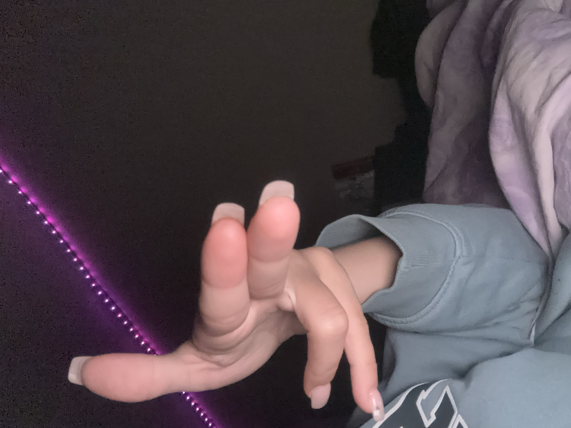 The image features a close-up perspective of a person's right hand and arm in a dimly lit indoor setting. The hand, adorned with long, light-colored (likely nude or light pink) acrylic or gel nails, is extended towards the viewer with fingers spread. The person is wearing a light blue or teal long-sleeved sweatshirt, with a portion of a white or light gray logo visible on the lower right of the garment, though illegible.
In the background, a string of purple LED lights runs diagonally across the left side of the frame, illuminating the dark environment. The upper right portion of the image shows folded fabric, possibly a blanket, in shades of purple or lavender, suggesting a bed or sofa. The overall lighting is dim, indicating it might be evening or night, with the purple LEDs providing the primary visible light source on the left. The scene focuses primarily on the hand and arm, with the surrounding details providing context to a casual, relaxed indoor environment. No specific activity beyond the hand's posture can be definitively identified.