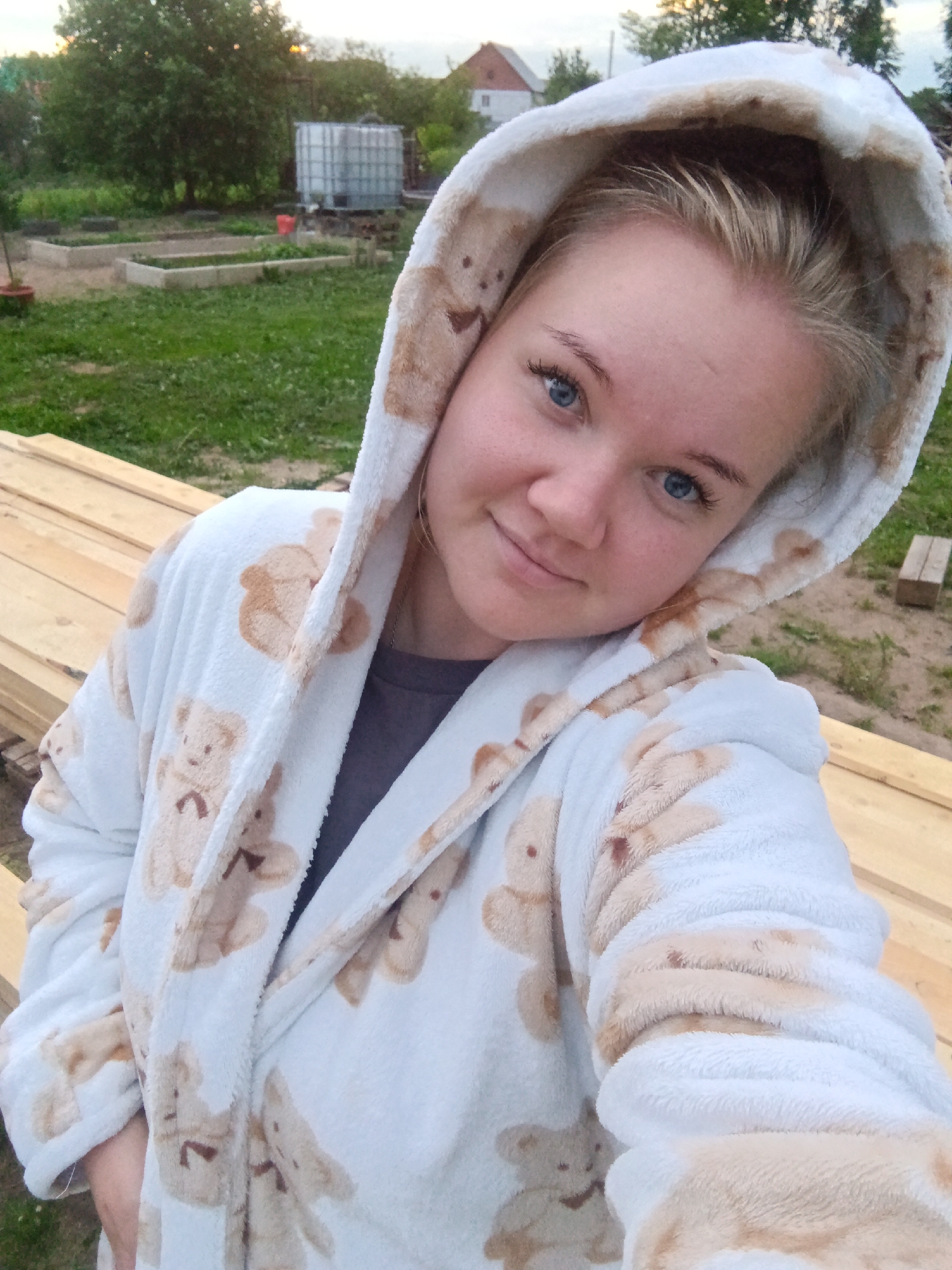 A young woman with blonde hair and blue eyes is taking a selfie outdoors. She is wearing a white hooded robe with a teddy bear pattern. The background shows a rural area with trees, grass, and some buildings, possibly in the city of Fokino, Russia. There are also some wooden planks in the foreground.