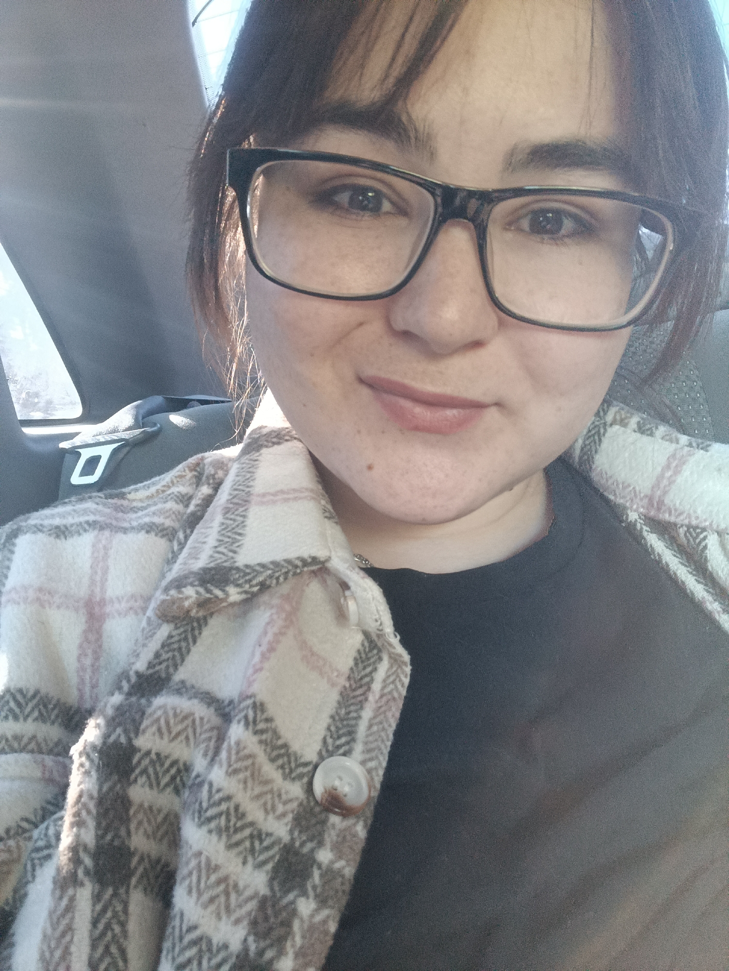 A close-up, chest-up selfie captures a young woman, likely in her 20s or early 30s, sitting inside a car. She has dark hair with bangs and wears black-framed eyeglasses. Her expression is pleasant, with a gentle, closed-mouth smile. Freckles are visible across her nose and cheeks.
She is wearing a black top beneath a cozy, plaid jacket or "shacket." The jacket features a pattern of light cream/white, pink, and gray/brown checks, with a noticeable herringbone texture woven into the fabric. A single, light brown button with a darker center is visible on the jacket's front. A thin silver necklace is also visible around her neck.
The setting is the interior of a car, indicated by a visible seatbelt buckle on the left side of the frame and the contours of car upholstery and a window. Bright, natural light streams in from the left, highlighting her hair and suggesting it is a sunny daytime. The overall impression is one of calm. No discernible text is visible in the image. The location context provided is Noviy, Russia.