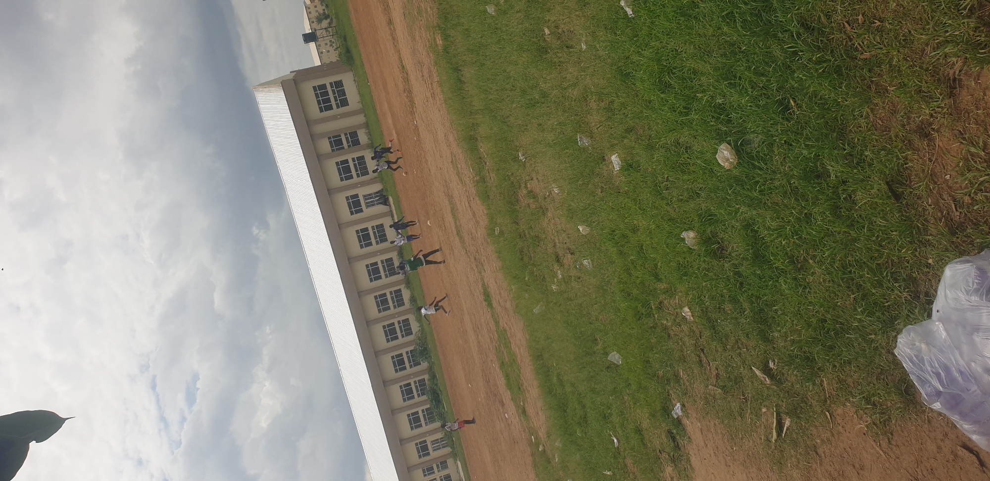 The image depicts an outdoor scene with a long, multi-story building running along the left side of the frame. The building has a modern, light-colored facade with numerous windows arranged in a grid pattern. A large, white, sloped roof extends from the building. To the right of the building is a dirt field, which transitions into a grassy area. Several people, appearing to be young men and boys, are scattered across the dirt field, some with what look like soccer balls at their feet. They are dressed in athletic wear, with some wearing white shirts and others green. The activity appears to be a casual game of soccer or a similar sport.
The ground is a mixture of bare earth and green grass, with scattered white plastic debris visible on the grass, suggesting some litter. The sky is overcast with white and gray clouds, indicating a potentially cloudy or partly cloudy day. The lighting suggests it could be daytime, possibly late morning or afternoon. The overall setting appears to be a school or community sports field in Yelwa, Nigeria, given the location context. There is no visible text in the image. The atmosphere seems relaxed and focused on the activity taking place.