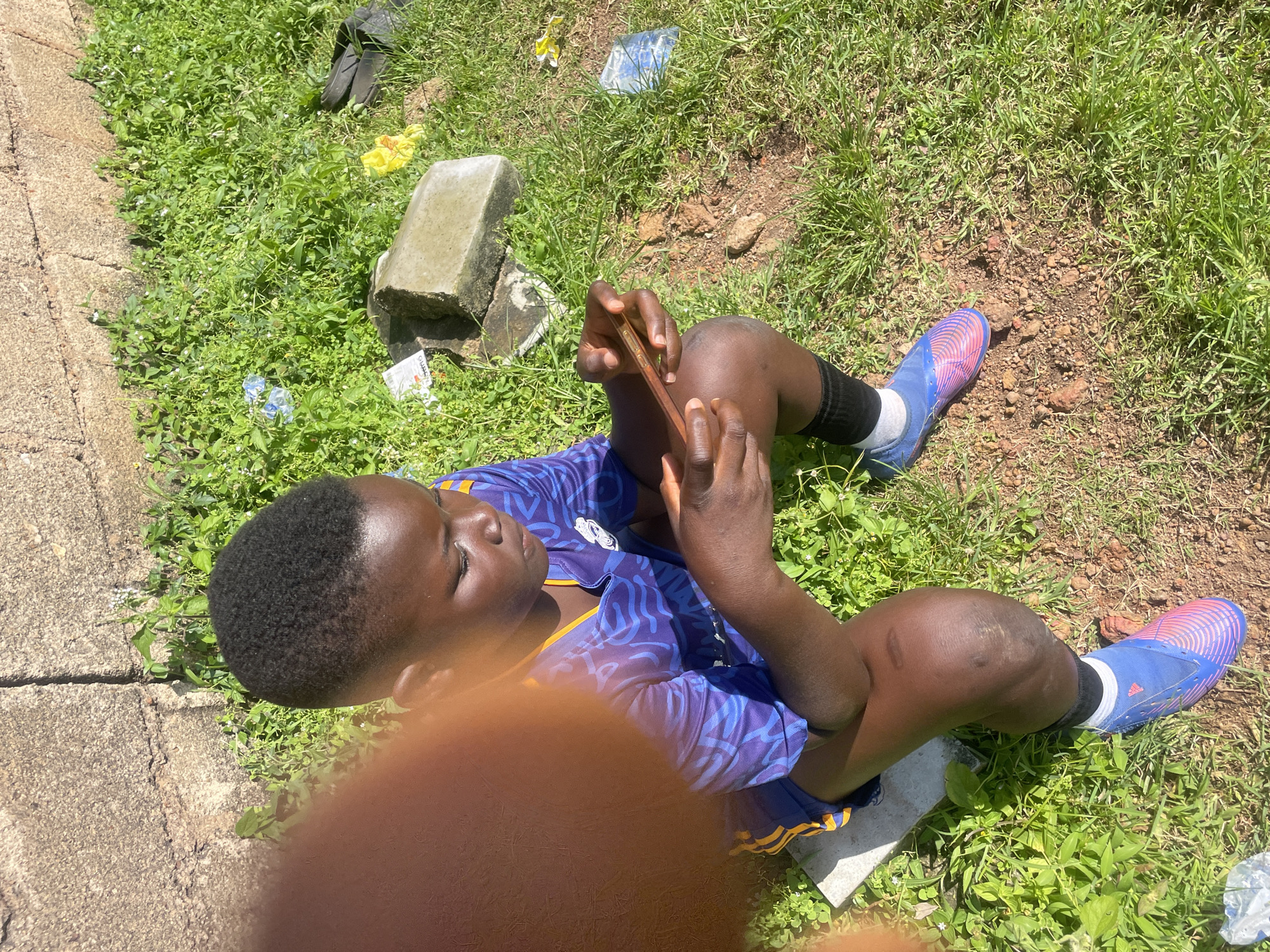 The image shows a young person sitting on the ground, focused on a smartphone. They are wearing a blue and purple soccer jersey with yellow accents and blue soccer cleats. The setting appears to be an outdoor area with green grass, dirt, and a rough, textured wall in the background, consistent with an urban environment in Jos, Nigeria. There is also some scattered debris, including plastic bags and what appears to be a concrete block, visible in the grass. The person's posture and grip on the phone suggest they are actively engaged with the device. A blurred orange shape in the foreground, likely a hand or arm, partially obscures the left side of the frame.
